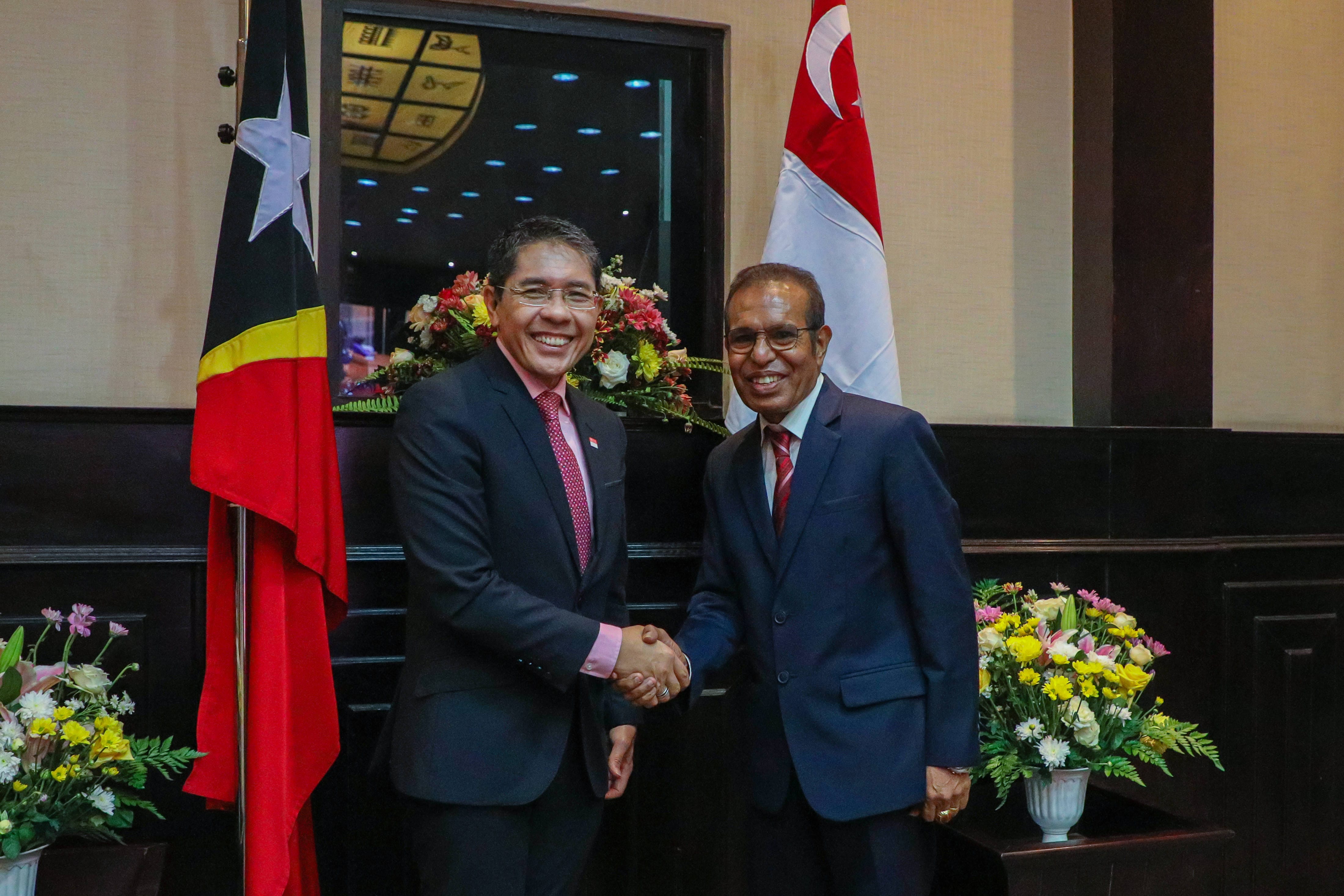Two men in suits shake hands, standing between flags of East Timor and Singapore.