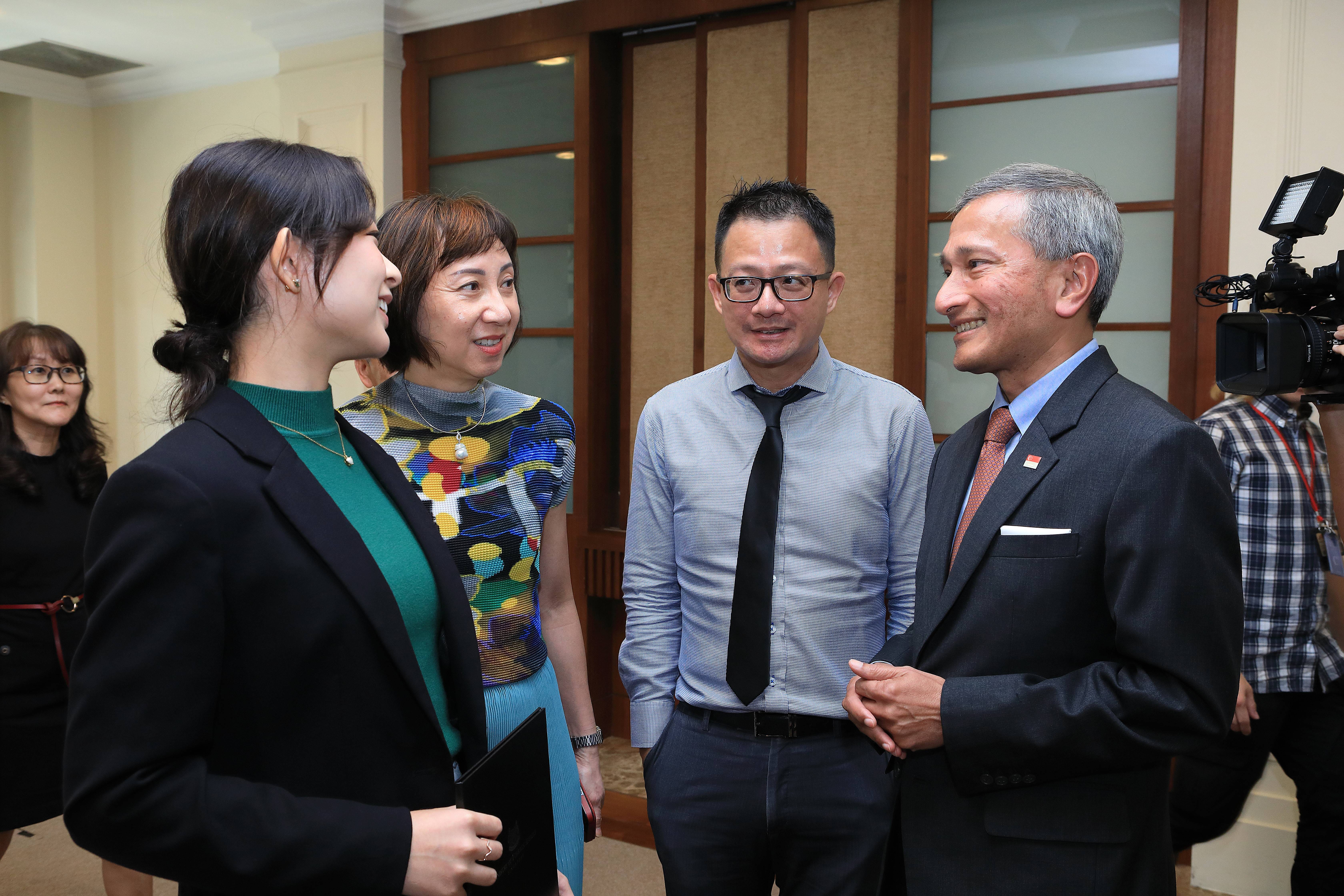 Group of four people in business attire converse; cameraman stands behind.