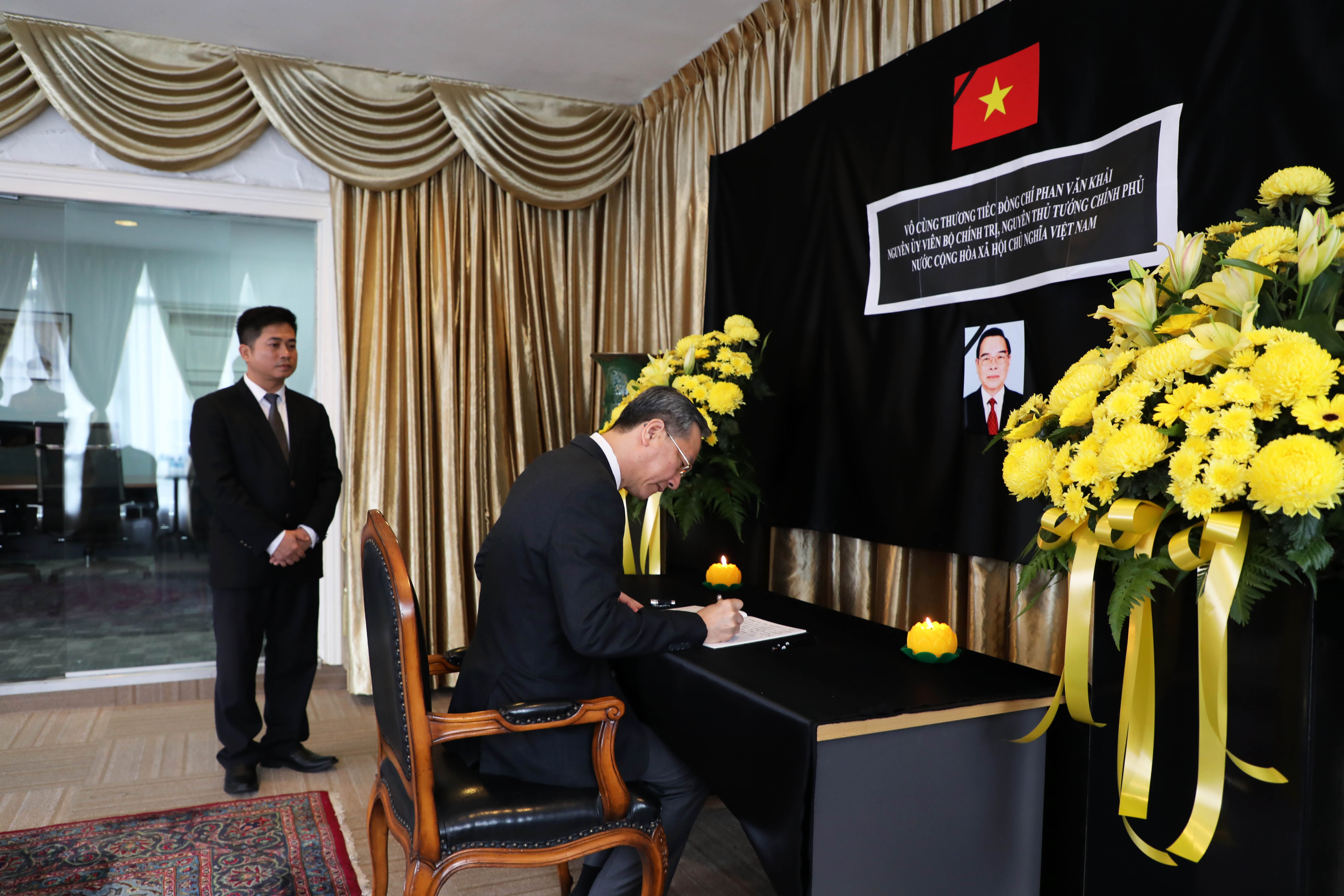 Man signs condolence book, beneath Vietnamese flag, portrait, and yellow flower arrangements.