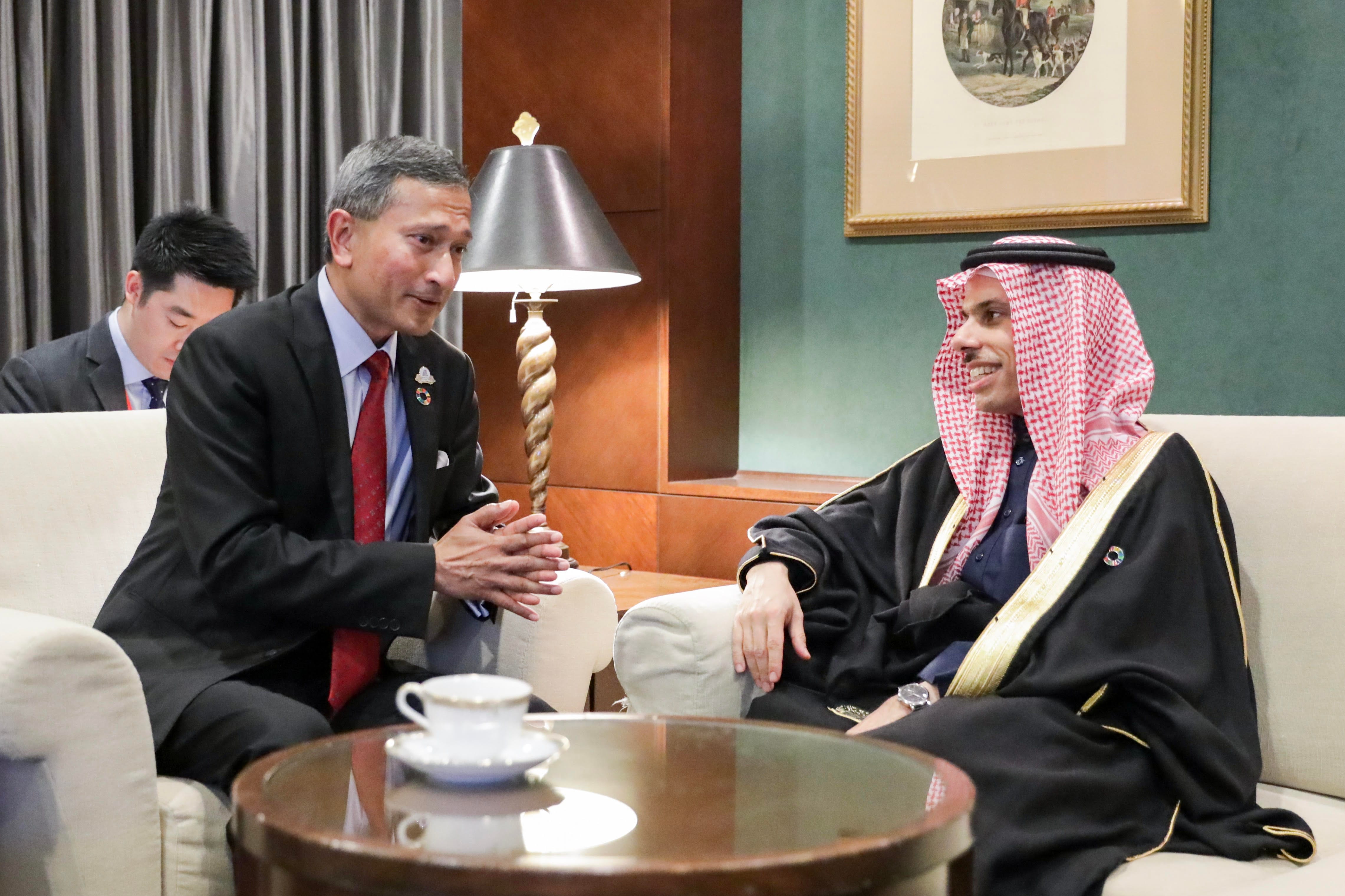 Two men in suits sit in armchairs, conversing near a coffee table with a teacup. One wears traditional Saudi clothing.