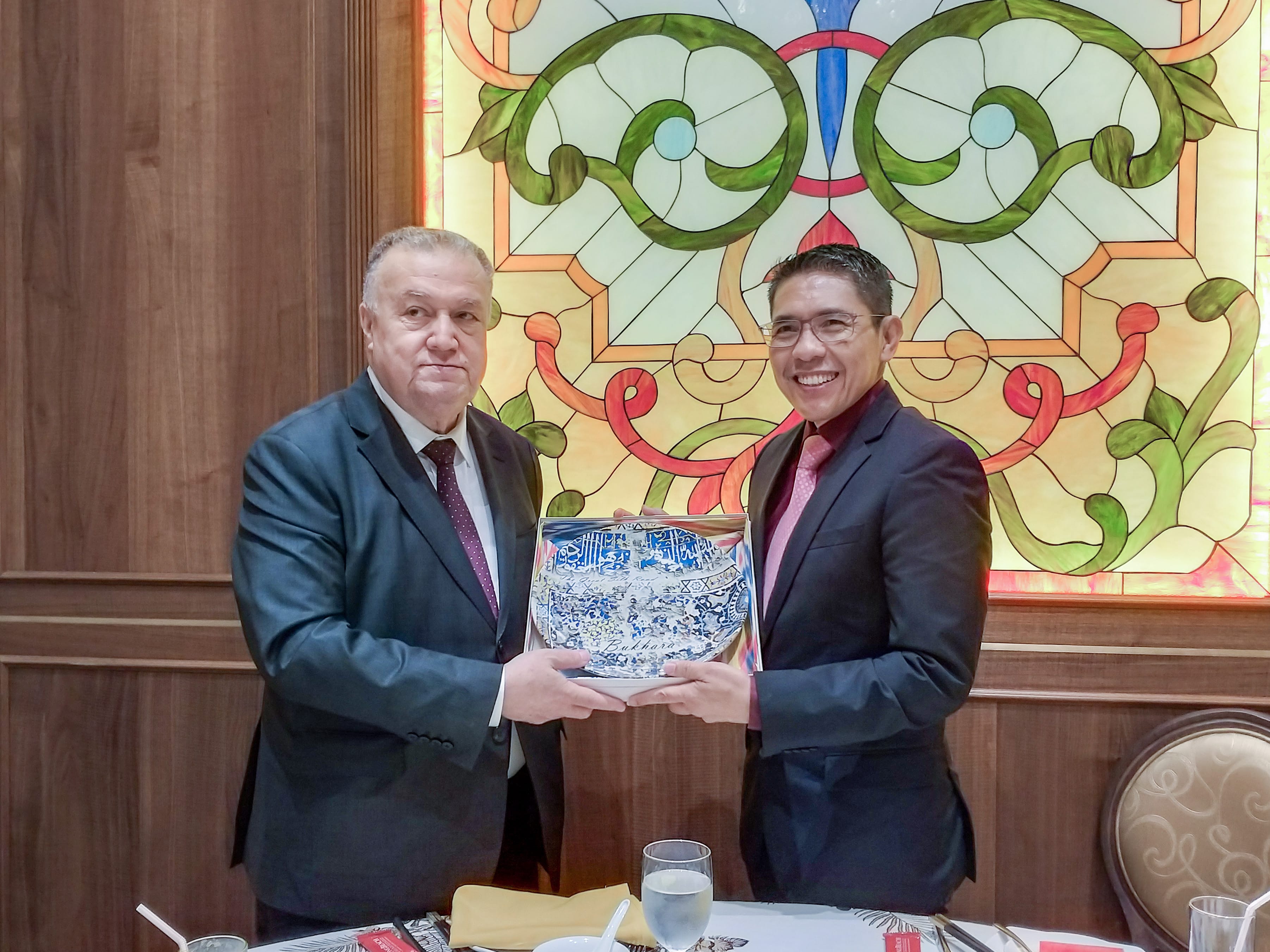 Two men holding a blue souvenir plate with 'Bukhara' text against a stained-glass backdrop.