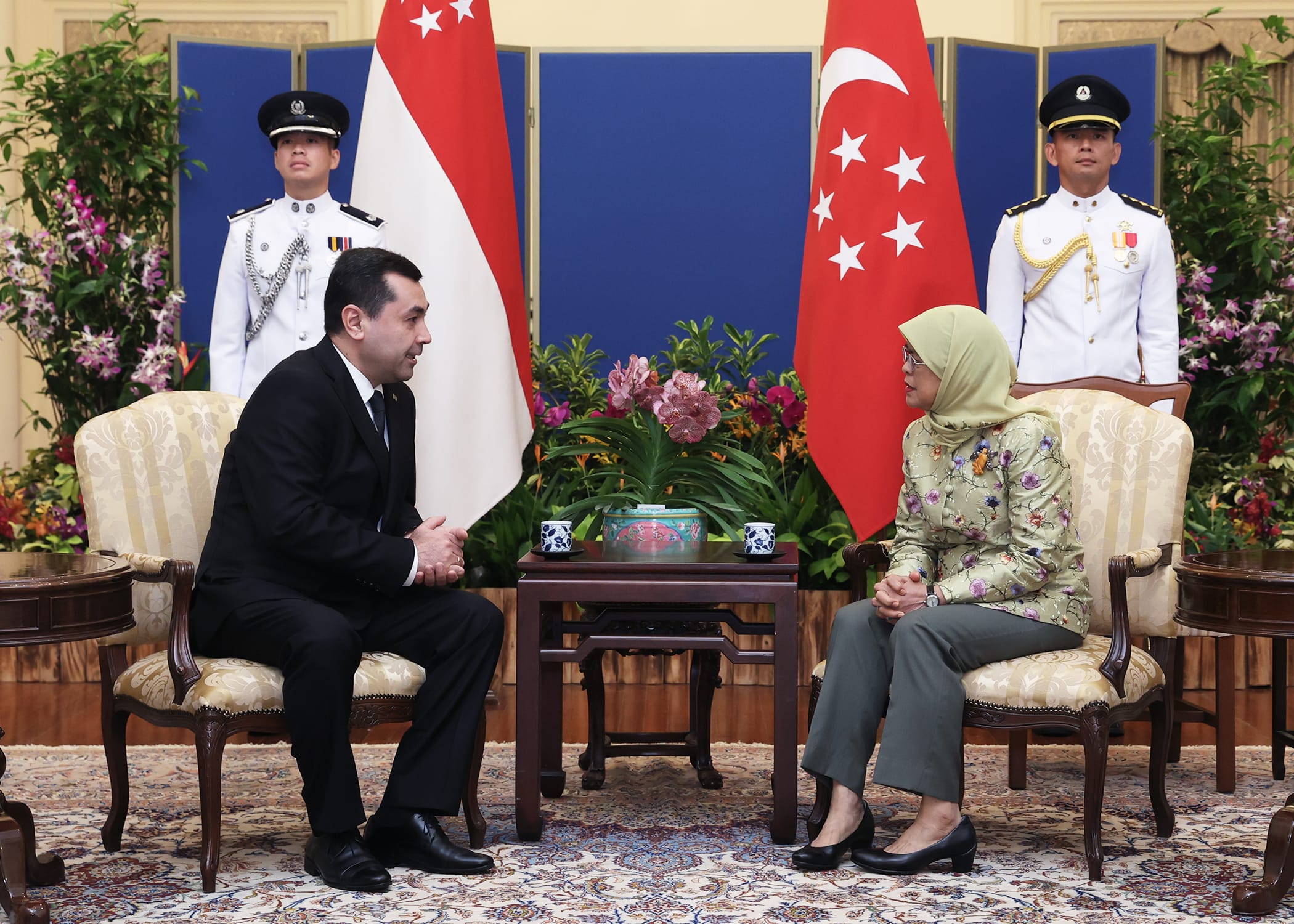 Man and woman seated across from each other, Singapore flags behind them, two guards stand at attention.