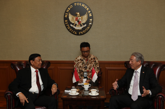 Two men in suits sit facing each other at a table with Indonesian and Singaporean flags. A man takes notes.