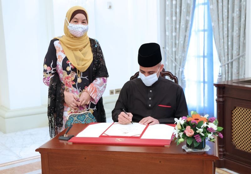 Man in black attire and mask signs document as woman in hijab looks on.