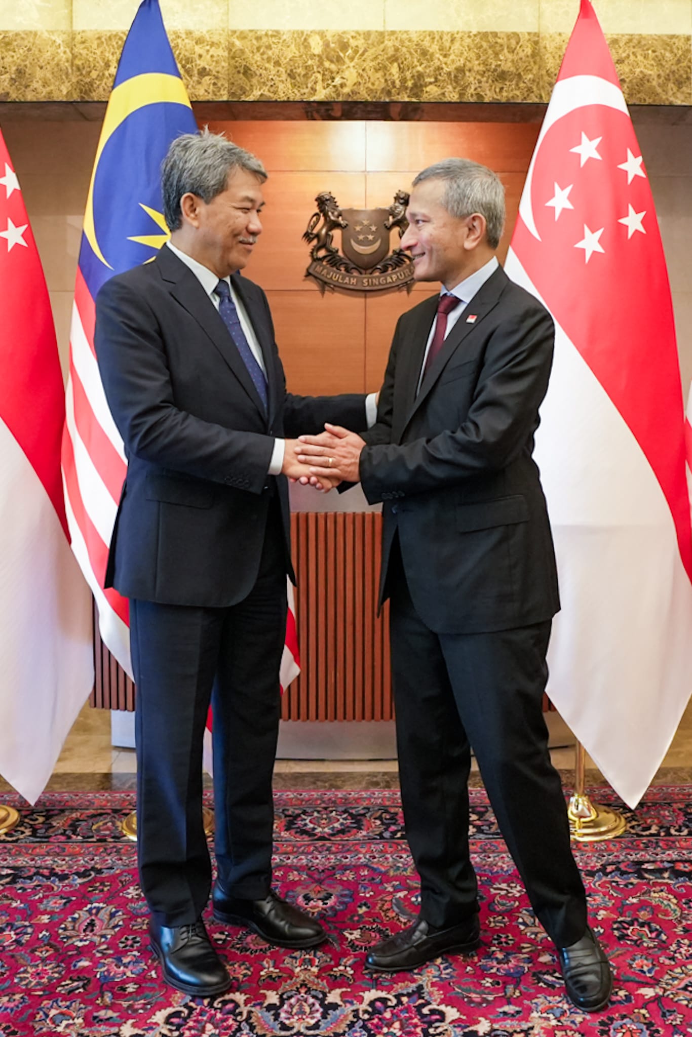 Two men in suits shake hands, with Malaysian and Singaporean flags behind them.