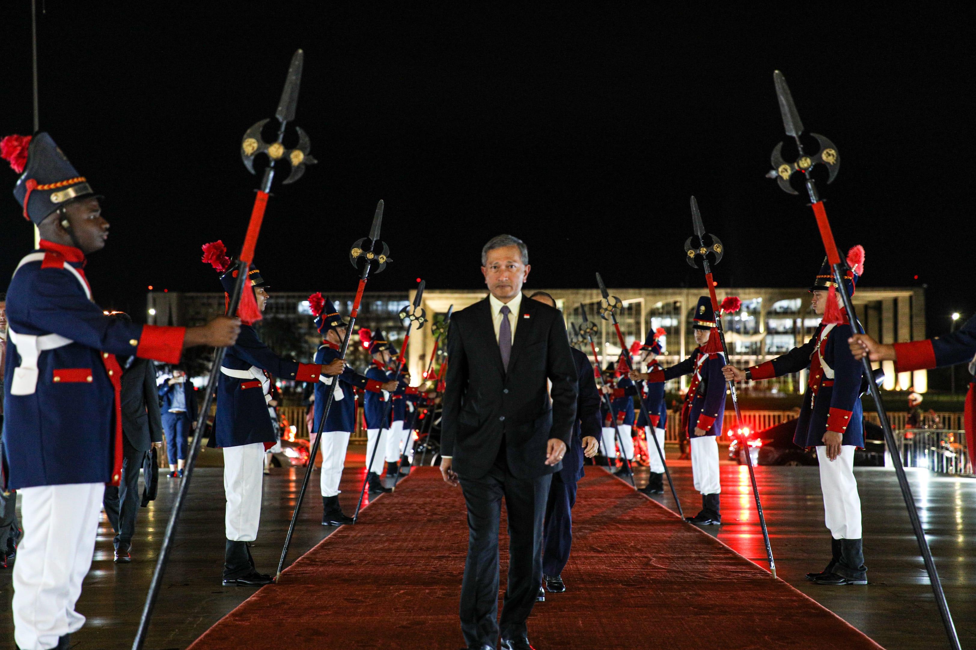 Man in suit walks red carpet flanked by honor guard holding spears, with a building in the background.