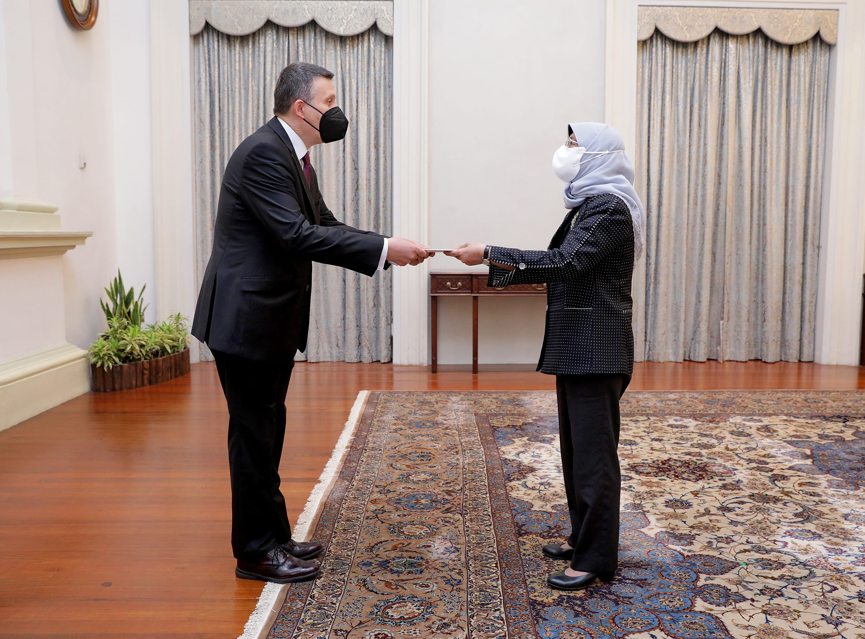 Masked man in suit presents a document to masked woman in hijab, indoors.