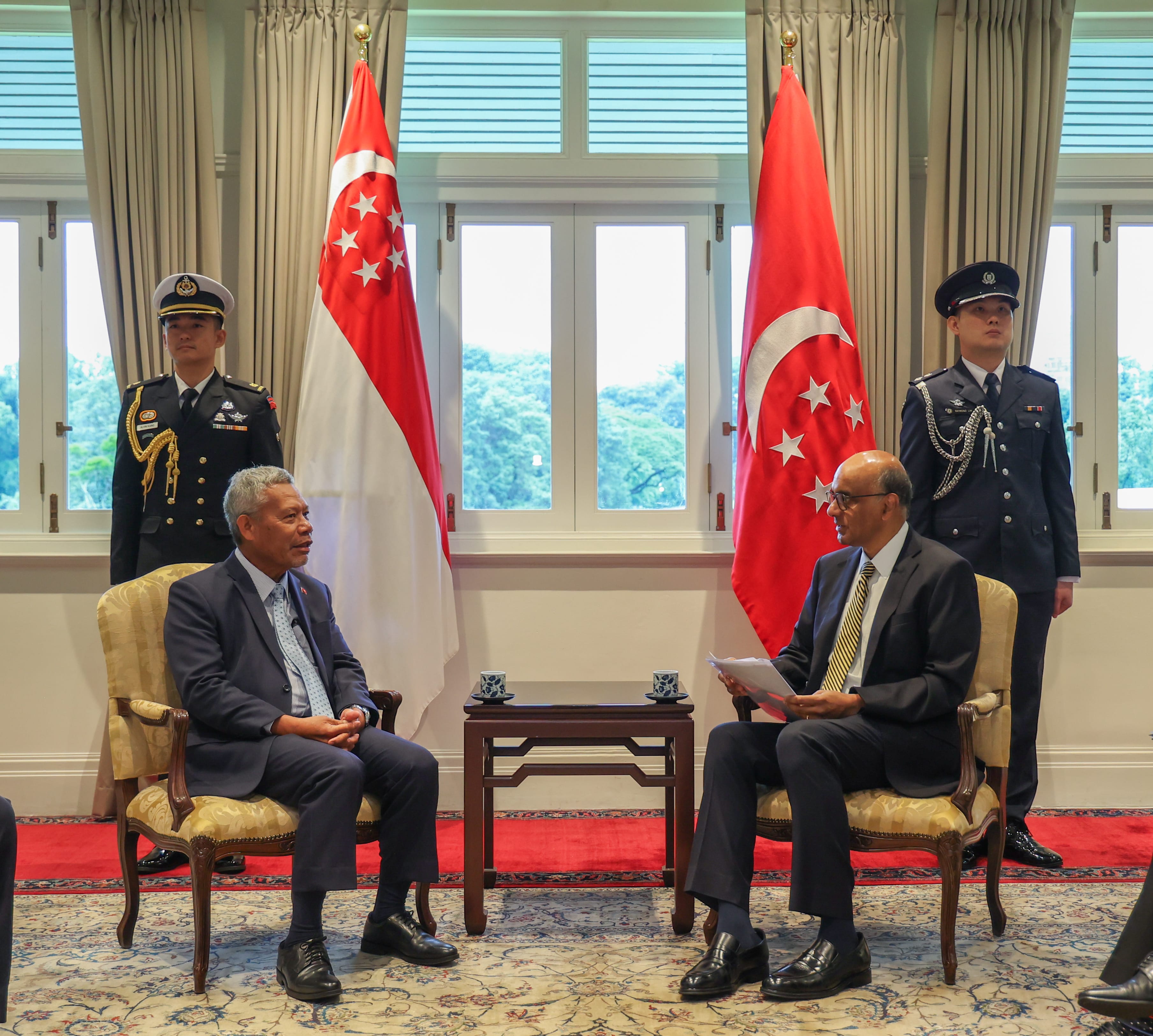 Two men in suits seated in chairs, Singapore flags, guards in uniform stand behind.