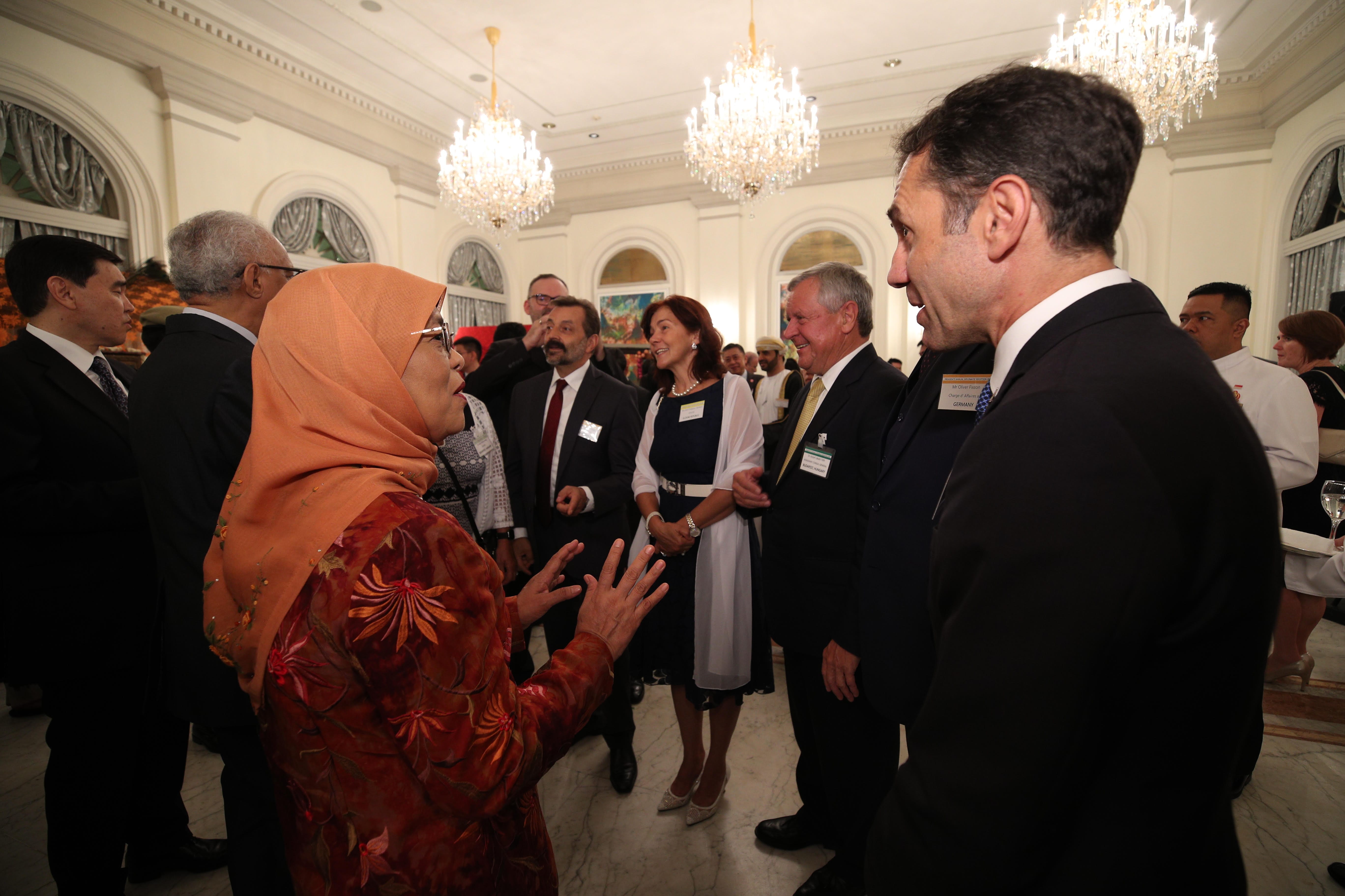 A group of people in formal attire converse in an ornate room with chandeliers.