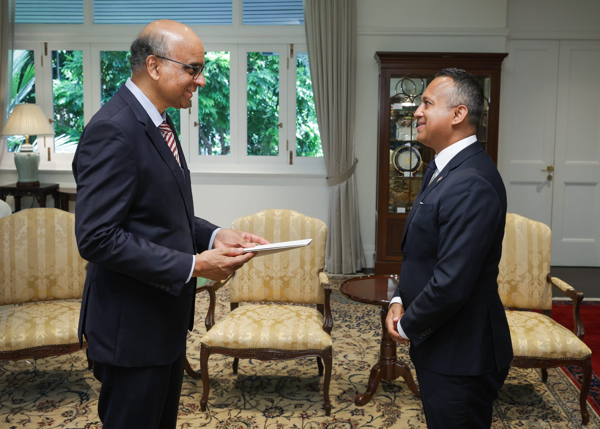 Two men in suits facing each other, one handing the other a document in an ornate room.
