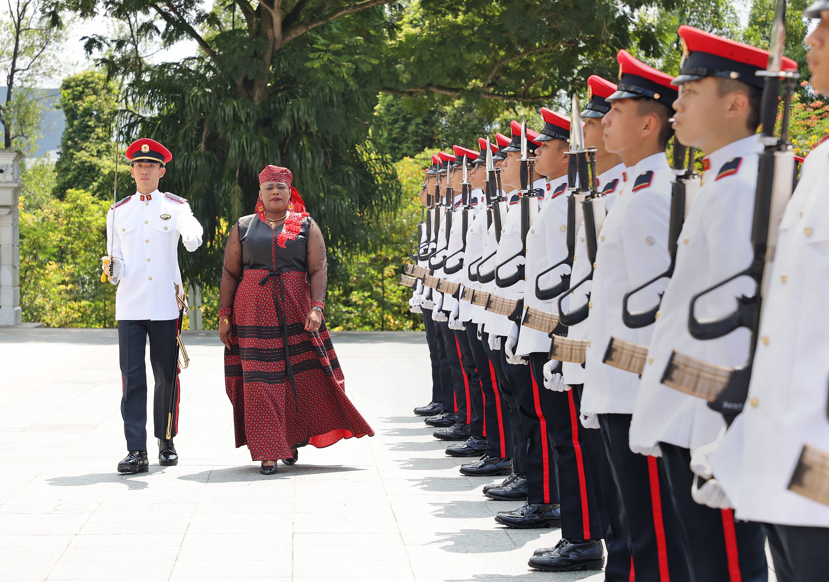 Woman in patterned dress inspects a military honor guard in white uniforms and red peaked caps.