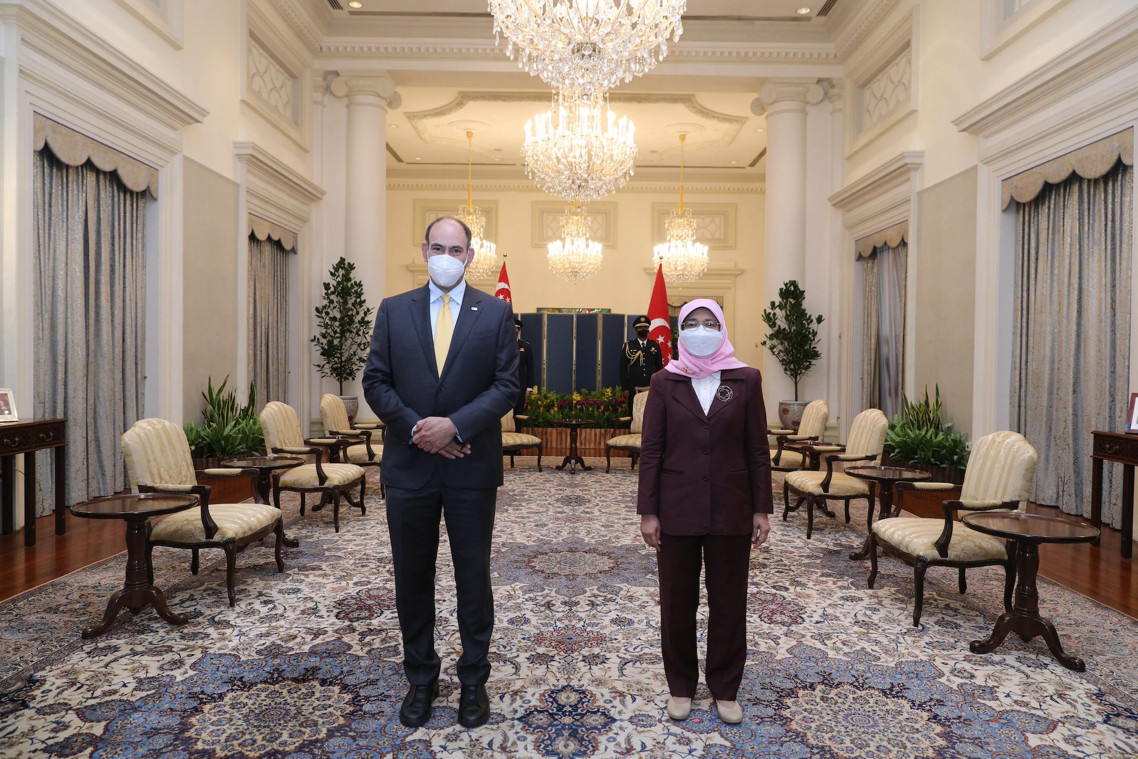 Two people in masks stand indoors with chandeliers, columns, flags and ornate carpeting.