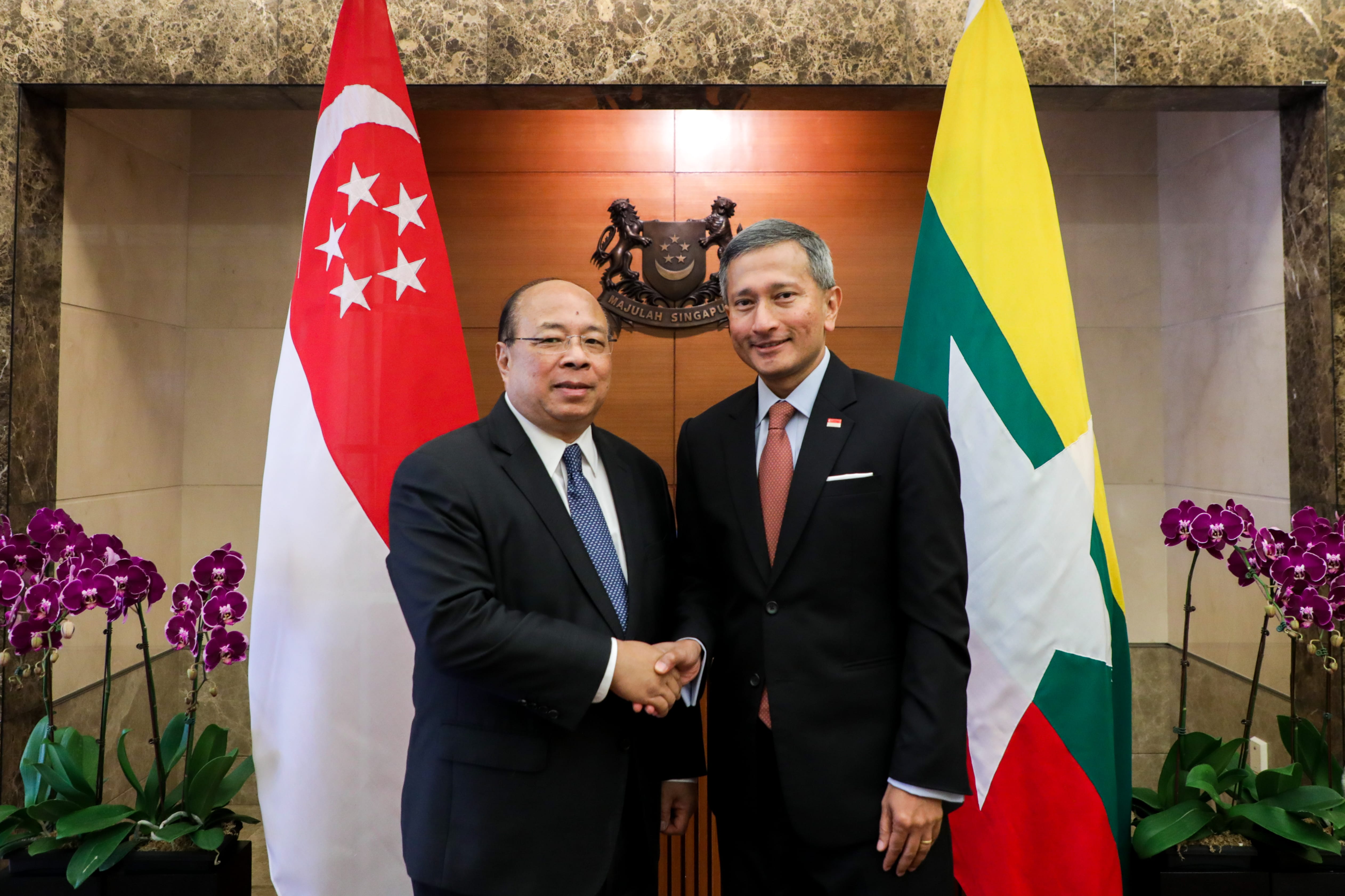 Two men in suits shaking hands between Singapore and Myanmar flags, and purple orchids.