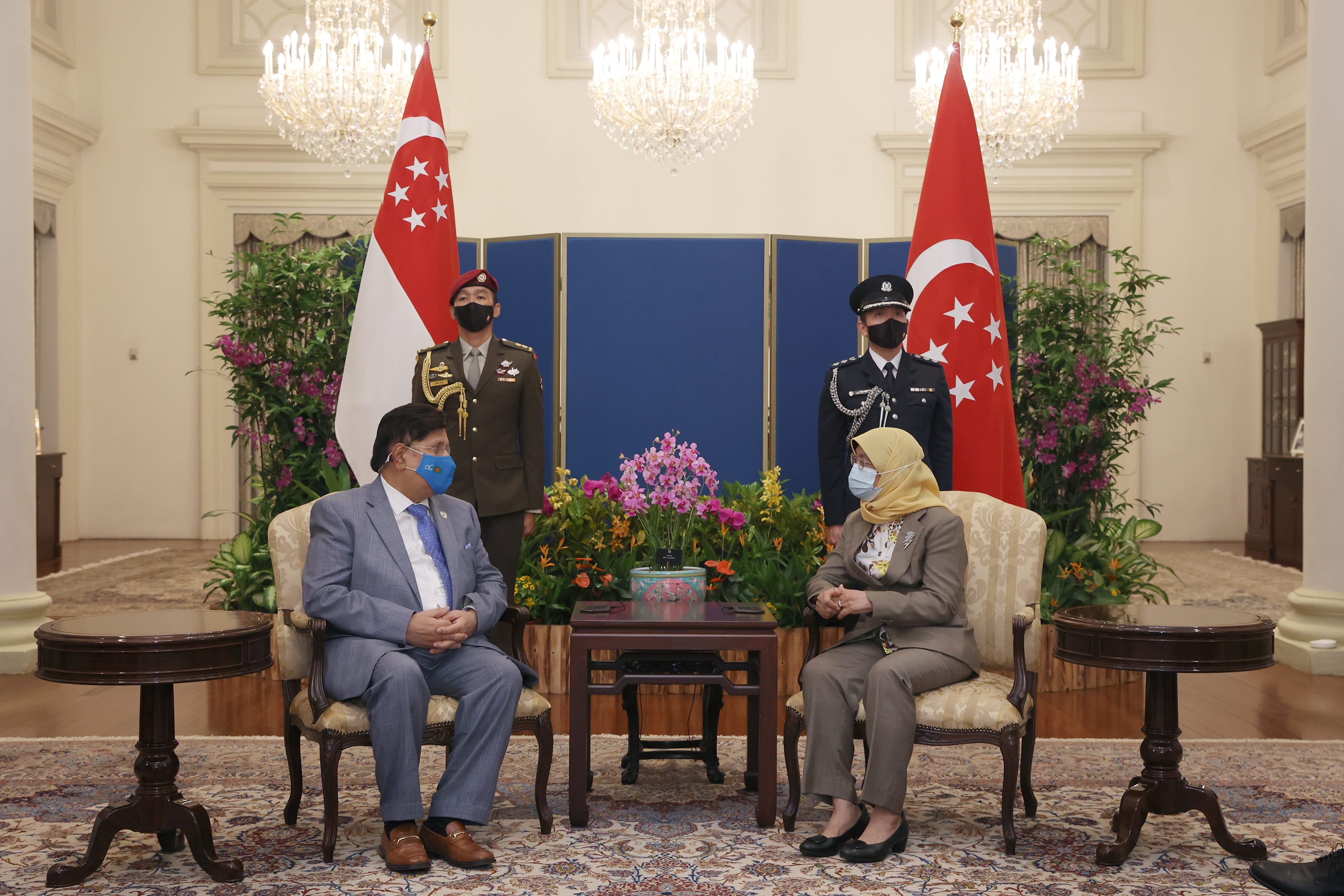 Halimah Yacob and foreign dignitary sit under chandeliers by Singapore flags and officers.