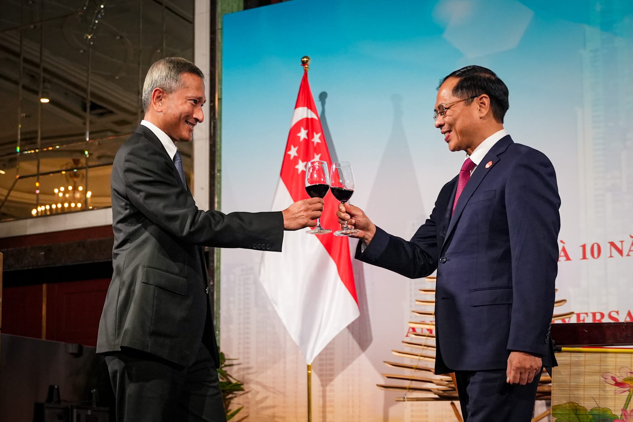 Two men in suits toasting red wine in front of the Singapore flag.