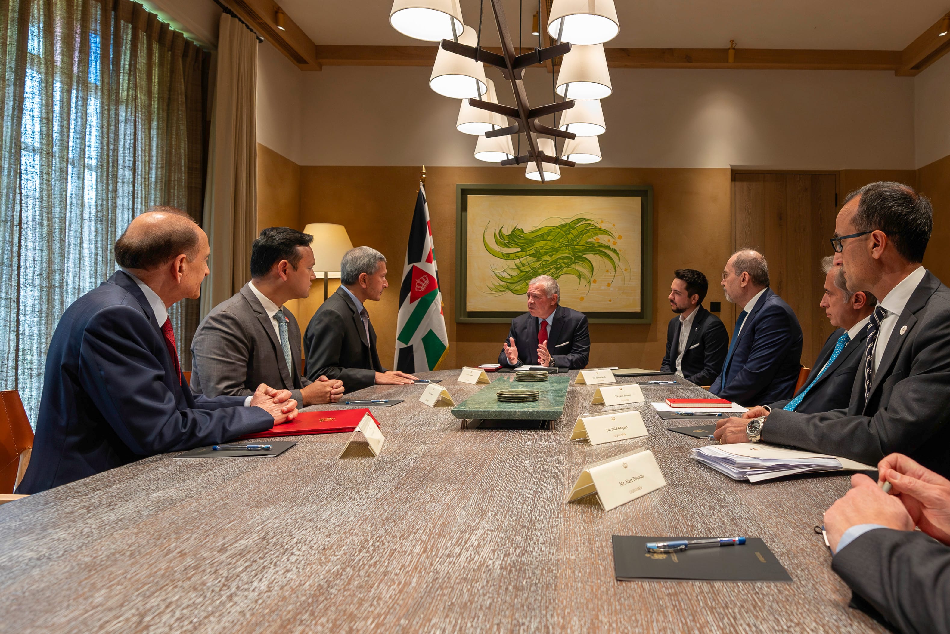 King Abdullah II at a long table meeting with several suited attendees. Jordan flag behind.