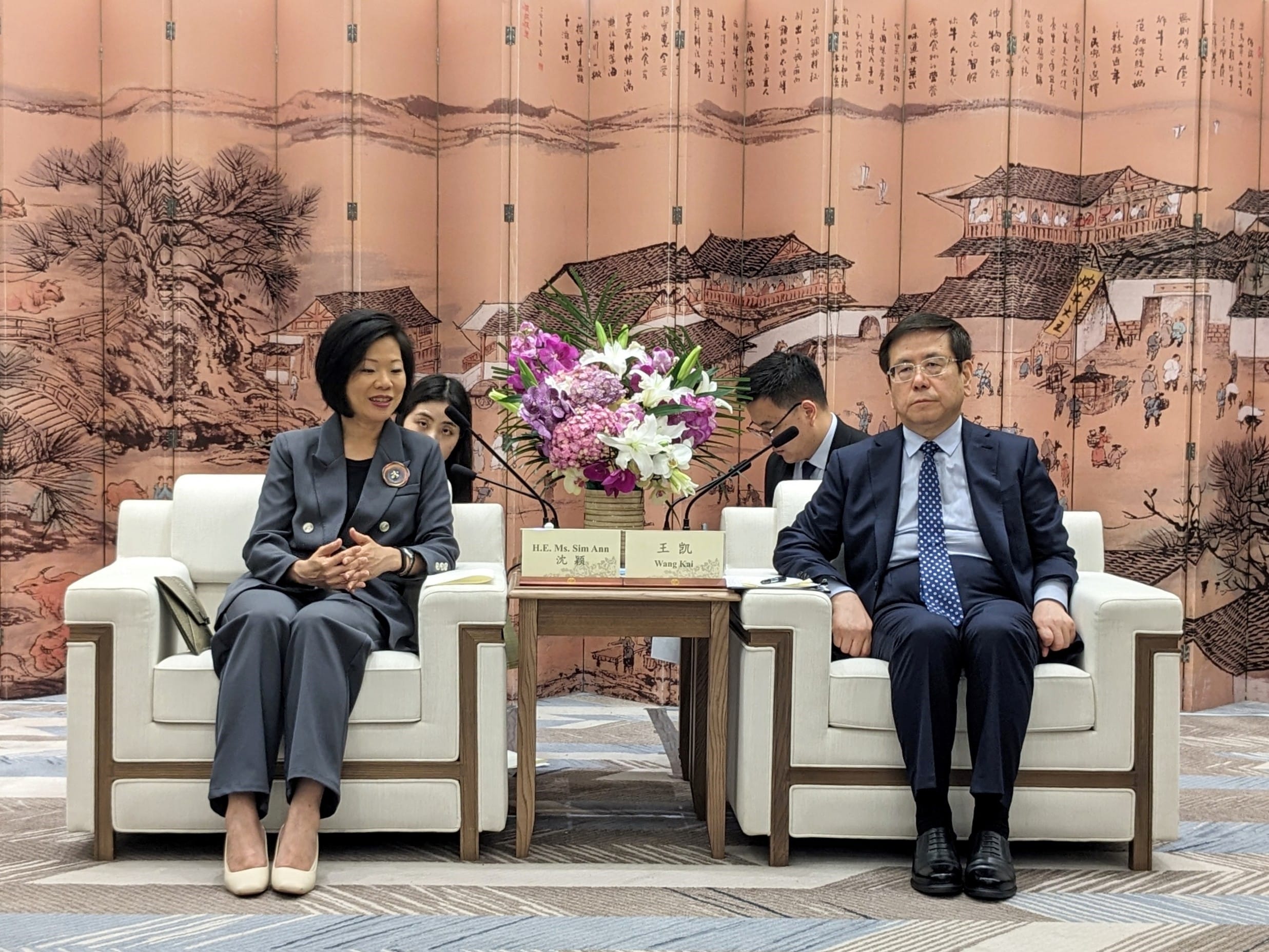 Three men in a room, Kuwaiti flag visible; framed portraits in background.