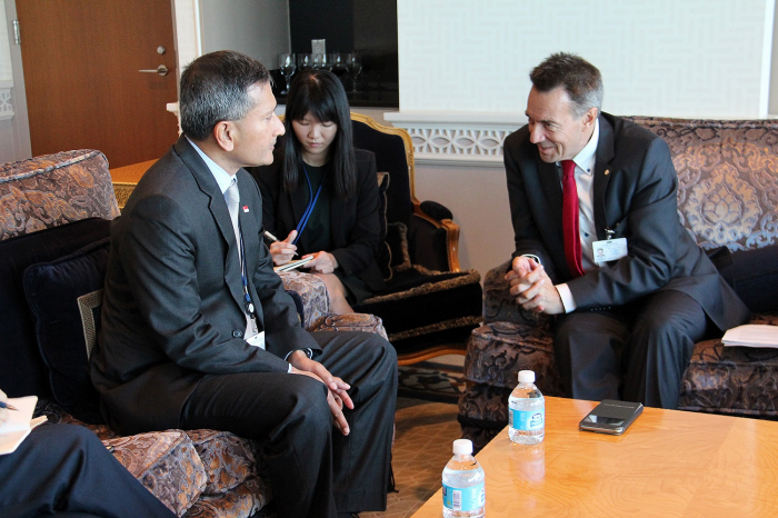 Two men in suits converse; woman takes notes. Water bottles & phone on table.