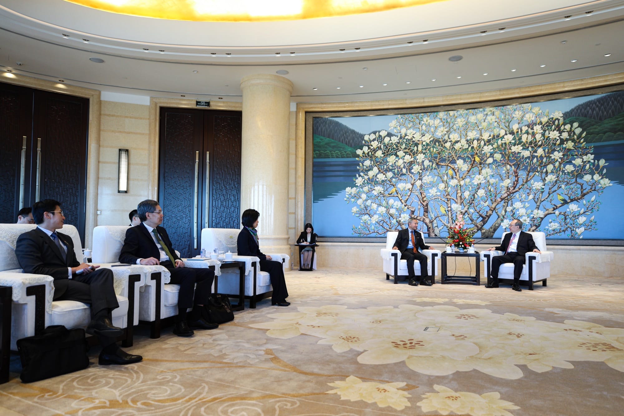 People in suits sit in white chairs in a large room with a mural.