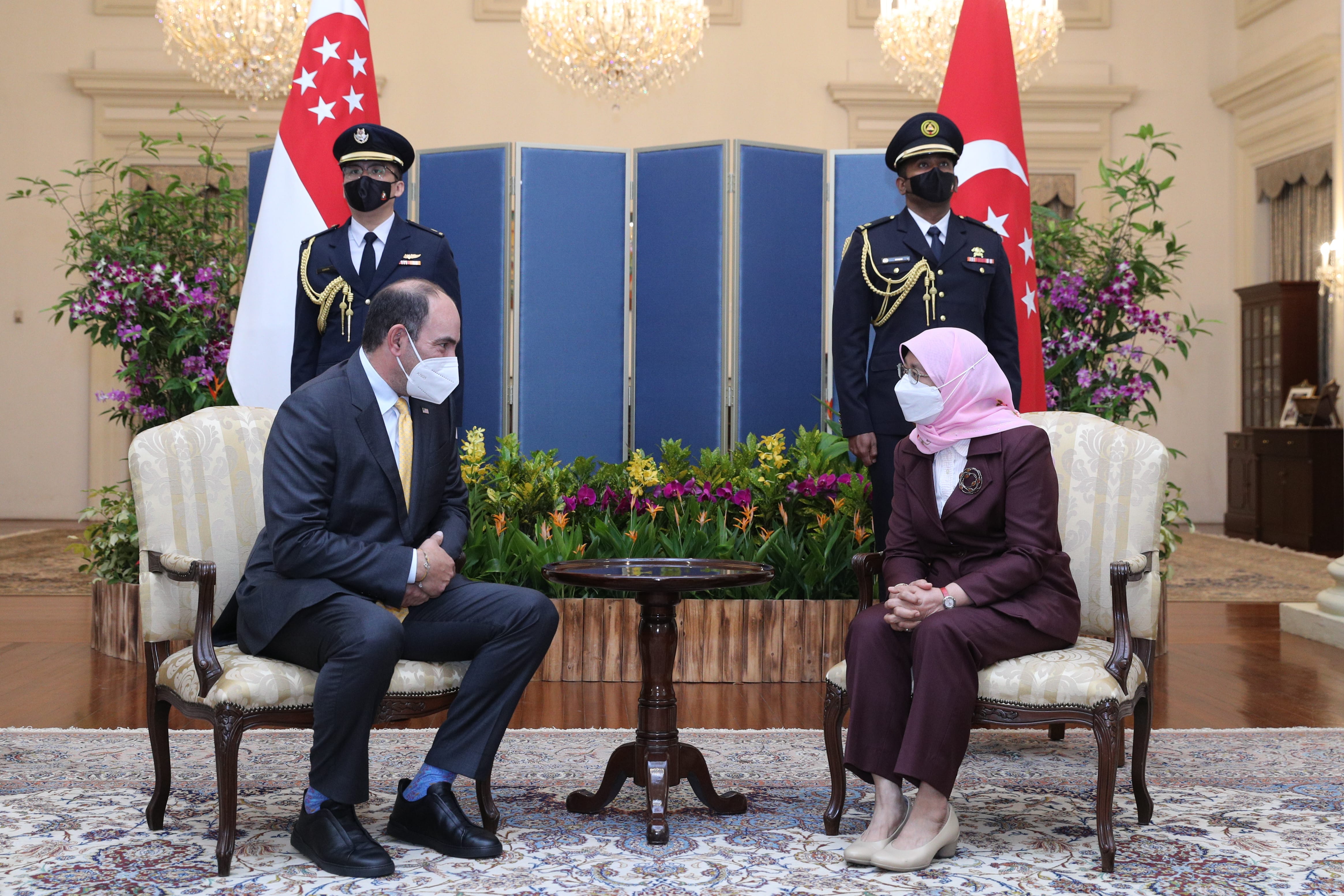Man and woman in suits seated, facing each other, with flags and uniformed guards in background. All wear masks.
