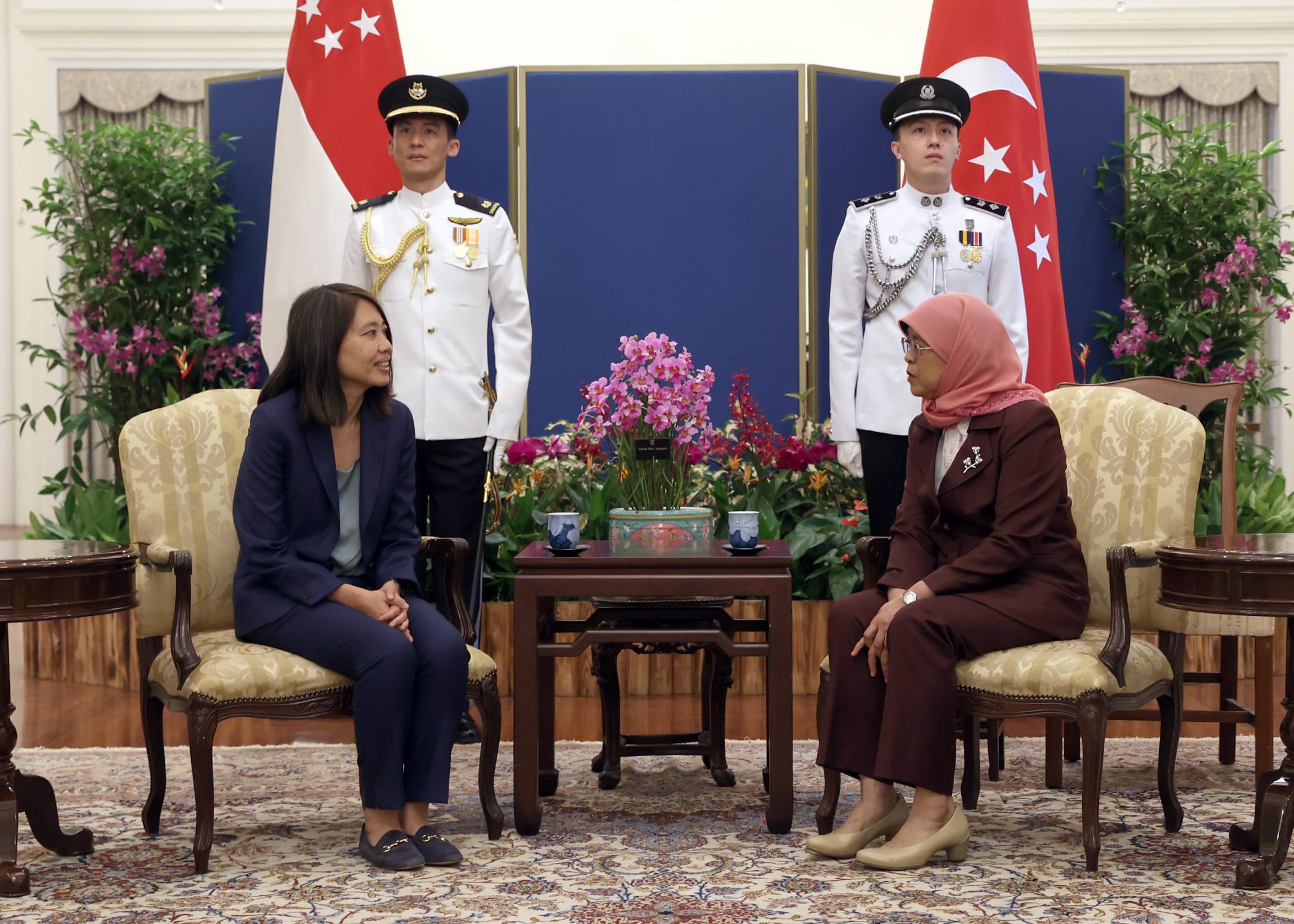 Two women in suits seated with guards and Singapore flags behind them.