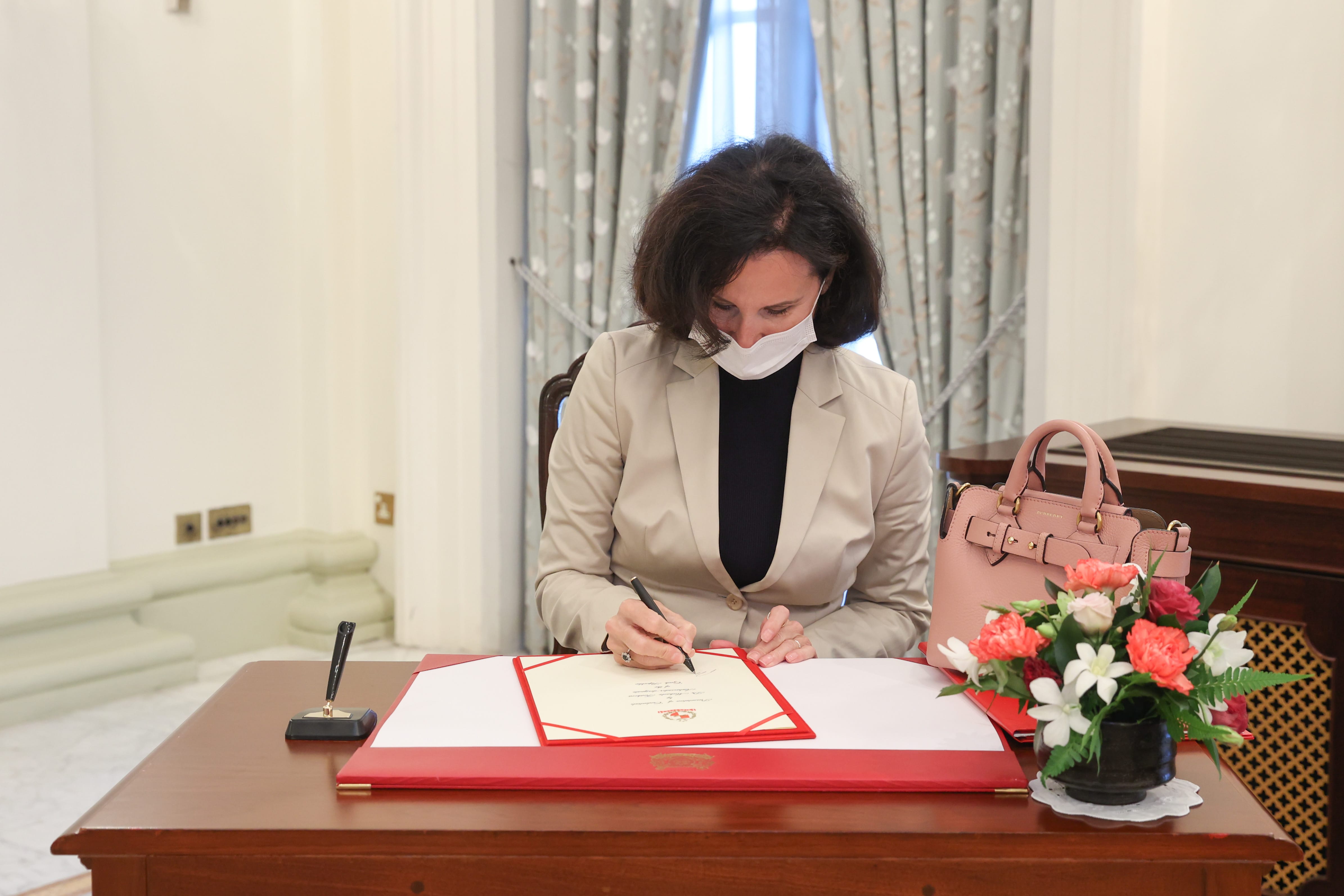 Woman in mask signs document on desk with flowers and Burberry handbag.