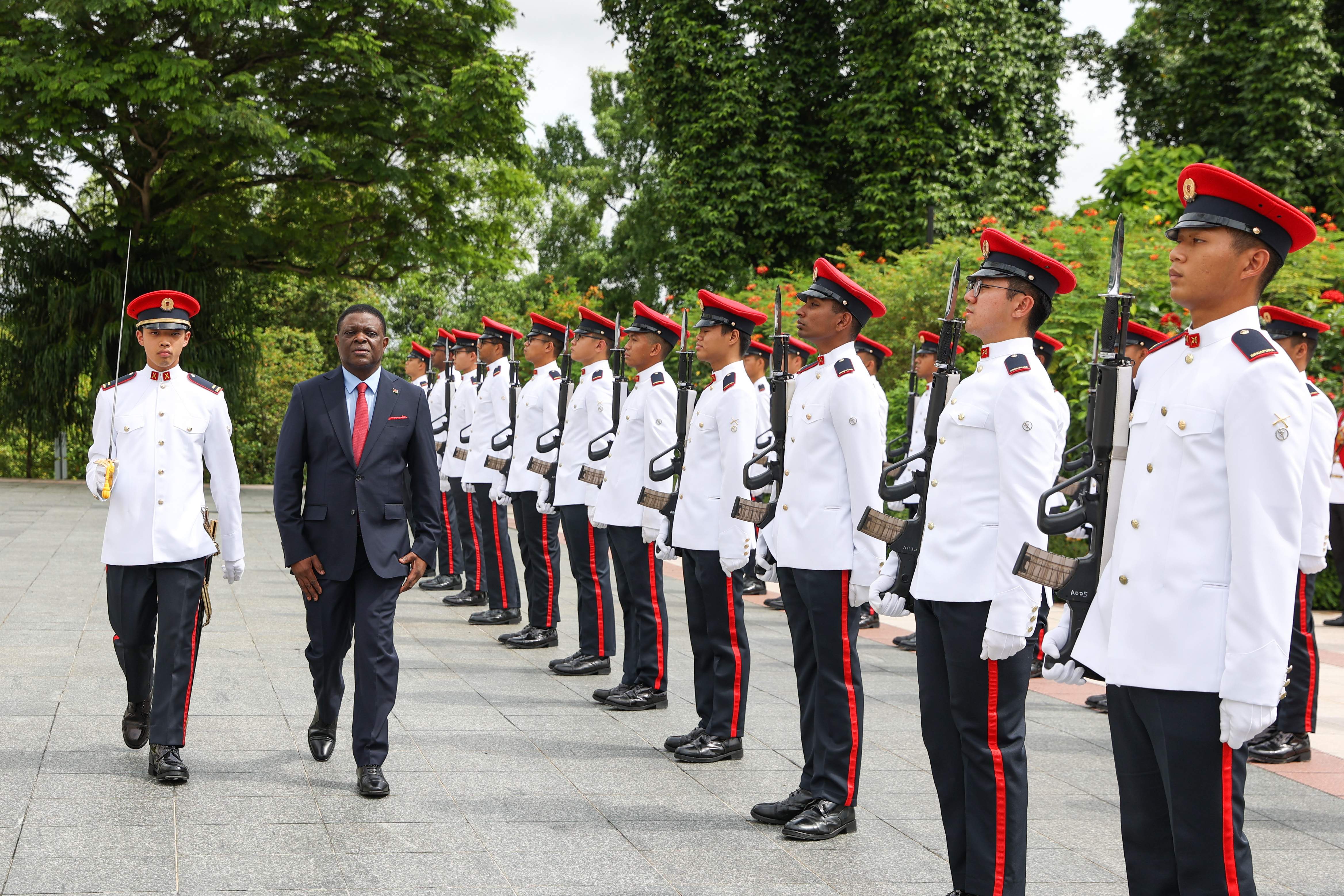 Man in suit walks past honor guard in white uniforms and red hats holding rifles.