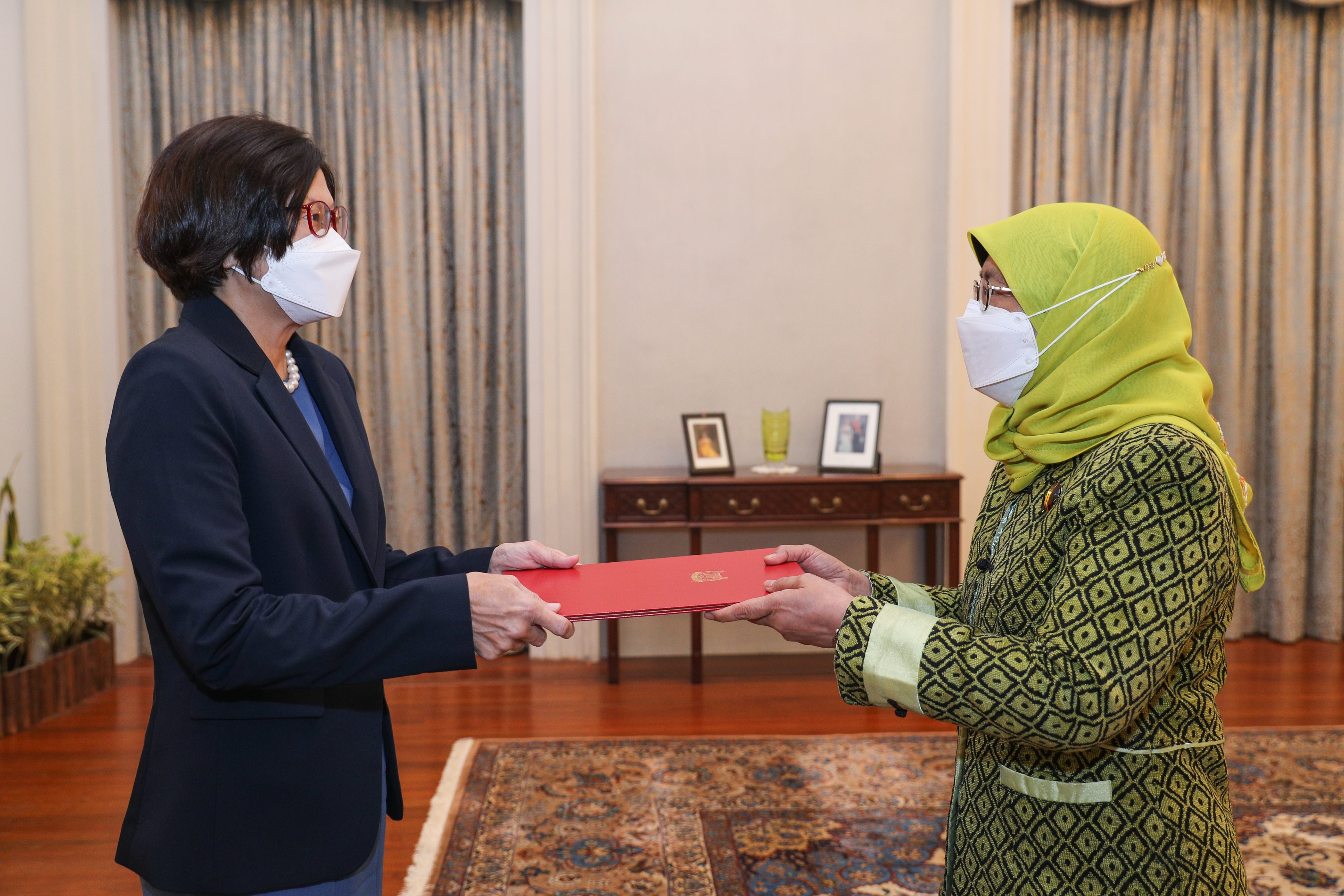 Two masked women exchange a red folder in a room with patterned carpet.