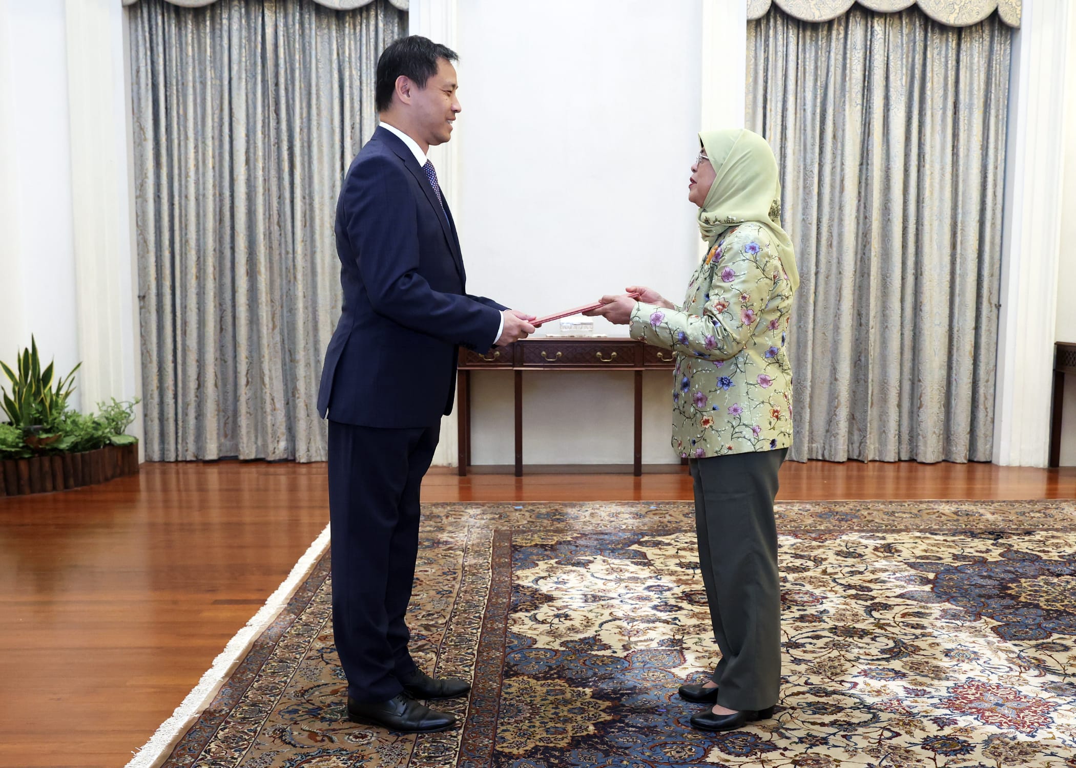 Two people exchanging a document. Man in suit, woman in hijab and floral jacket.