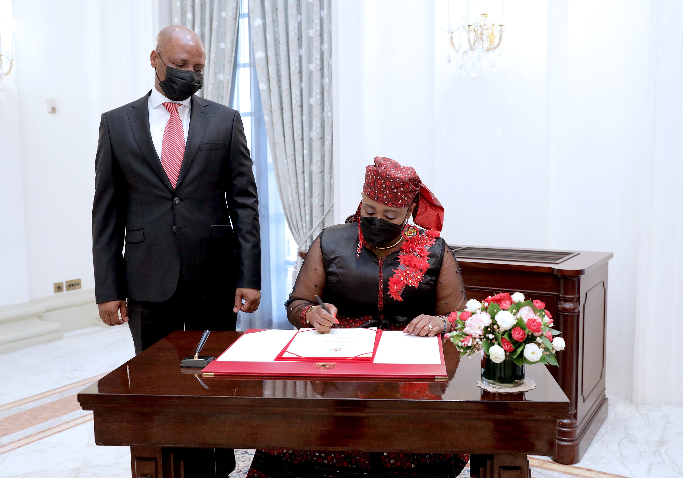 Woman signs document at a desk with a man standing nearby, both wearing face masks.