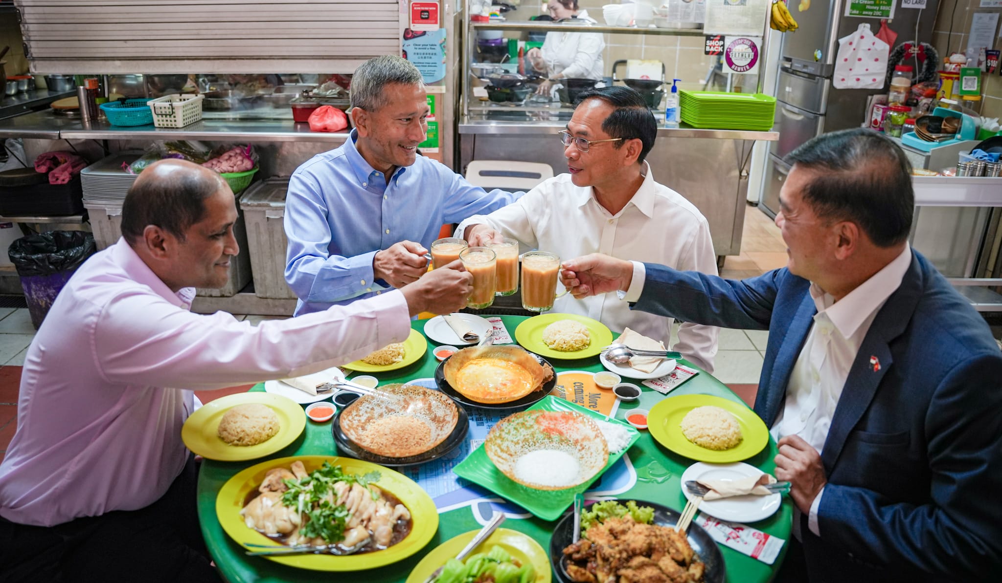 Four men in shirts clink glasses of tea at a table laden with food dishes.
