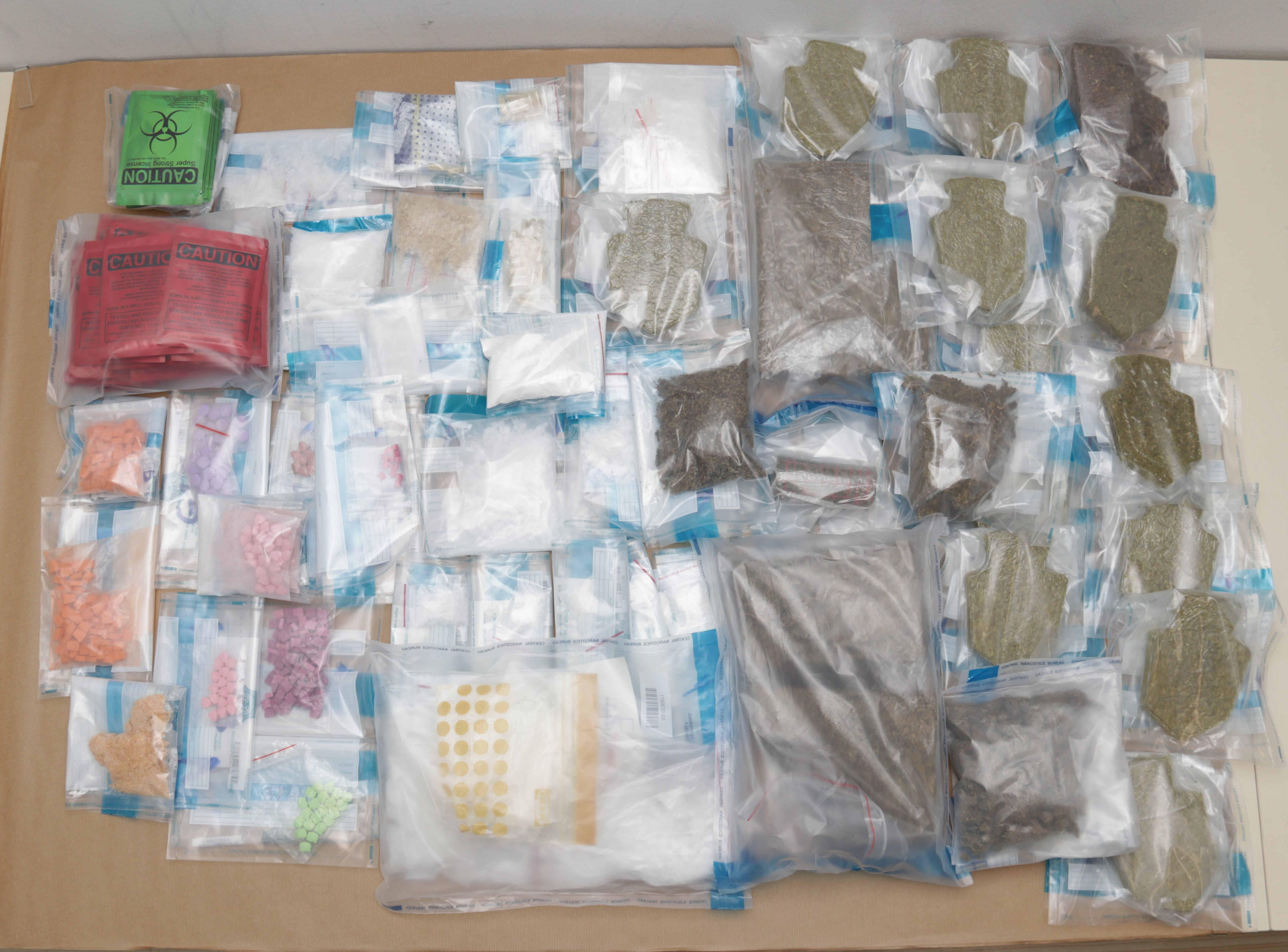 Multiple plastic bags of pills, powders, and plant matter, some labeled "CAUTION" or with biohazard symbols, on a surface.