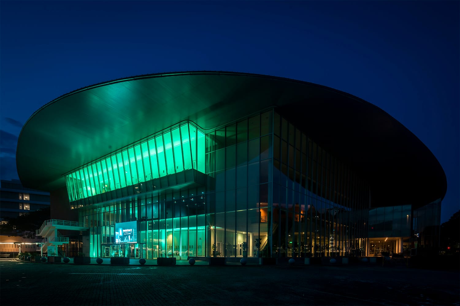 Modern glass building illuminated in green, with a large curved roof at night.