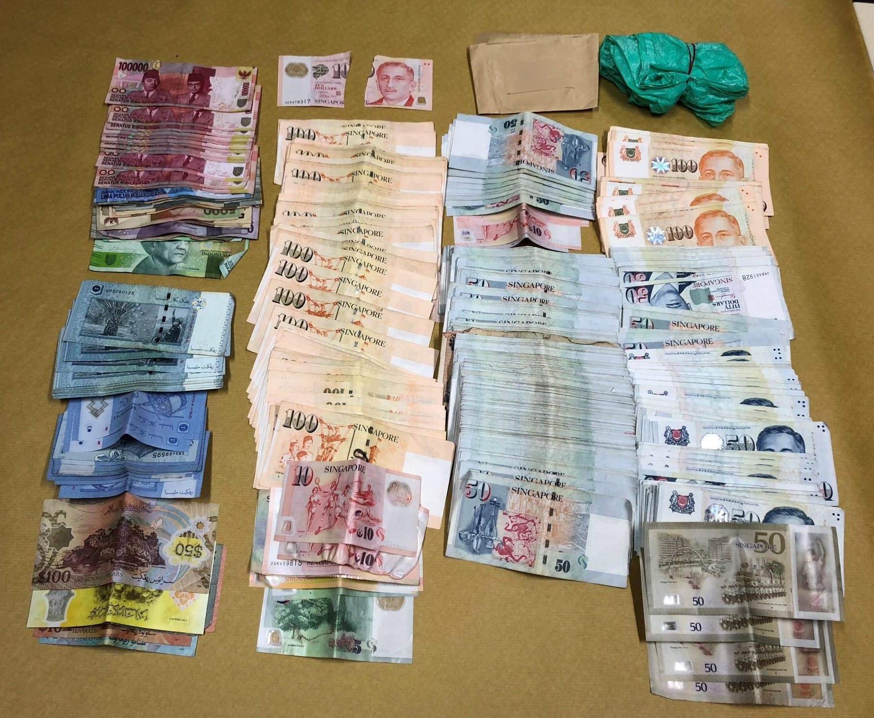 Stacks of Singapore, Malaysian, Indonesian currency, and two green plastic bags on tan surface.
