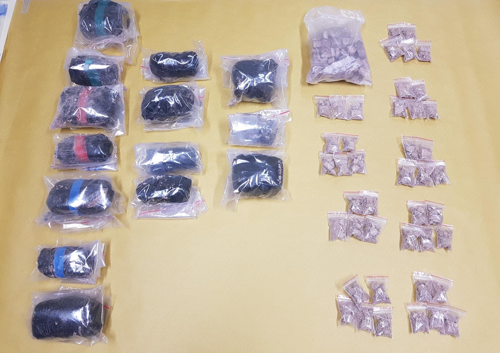 Packets of dark, rock-like material, some marked with colored tape, and smaller bags of powder on a yellow surface.