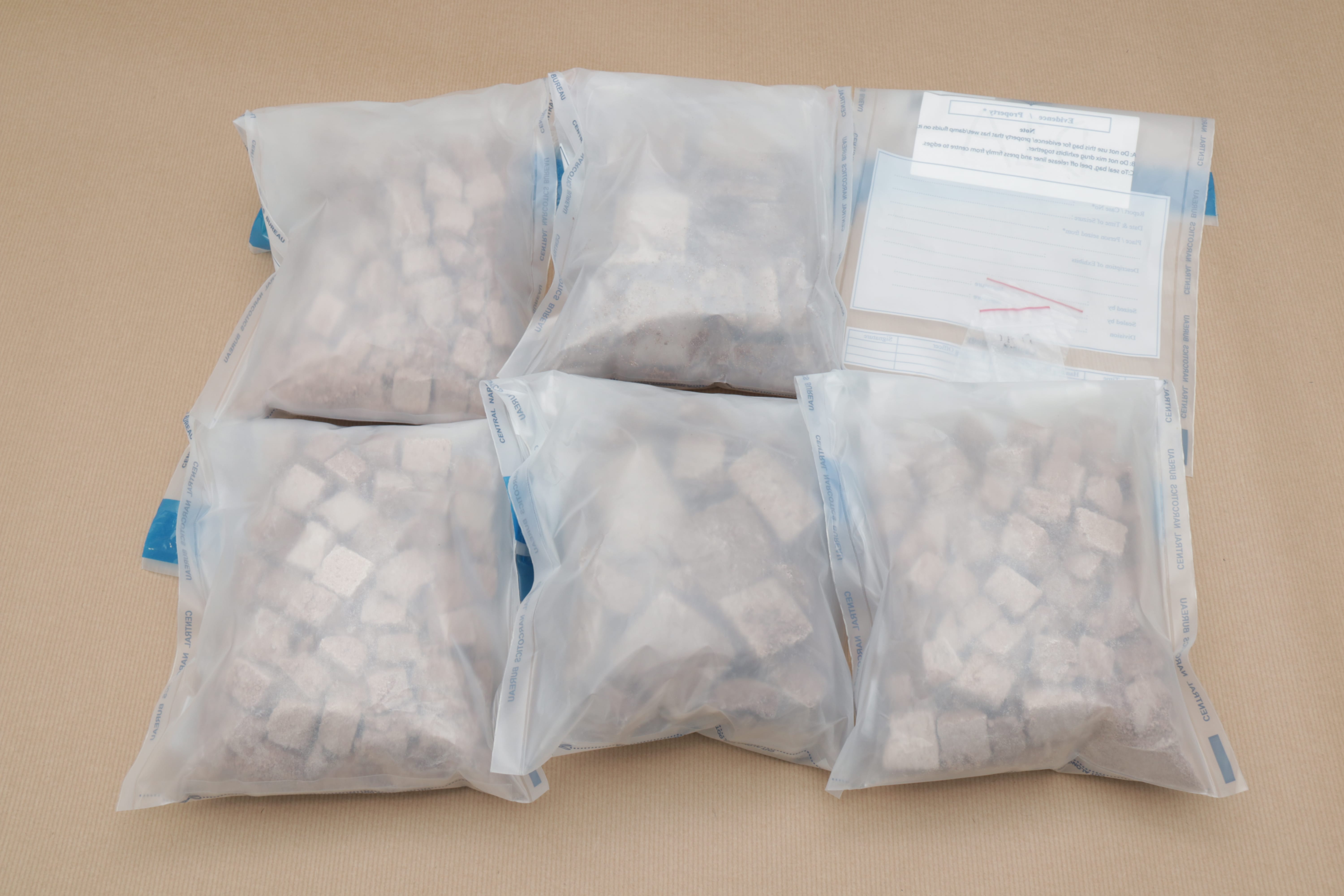 Five evidence bags filled with gray cube-shaped items and evidence labels, on a tan surface.