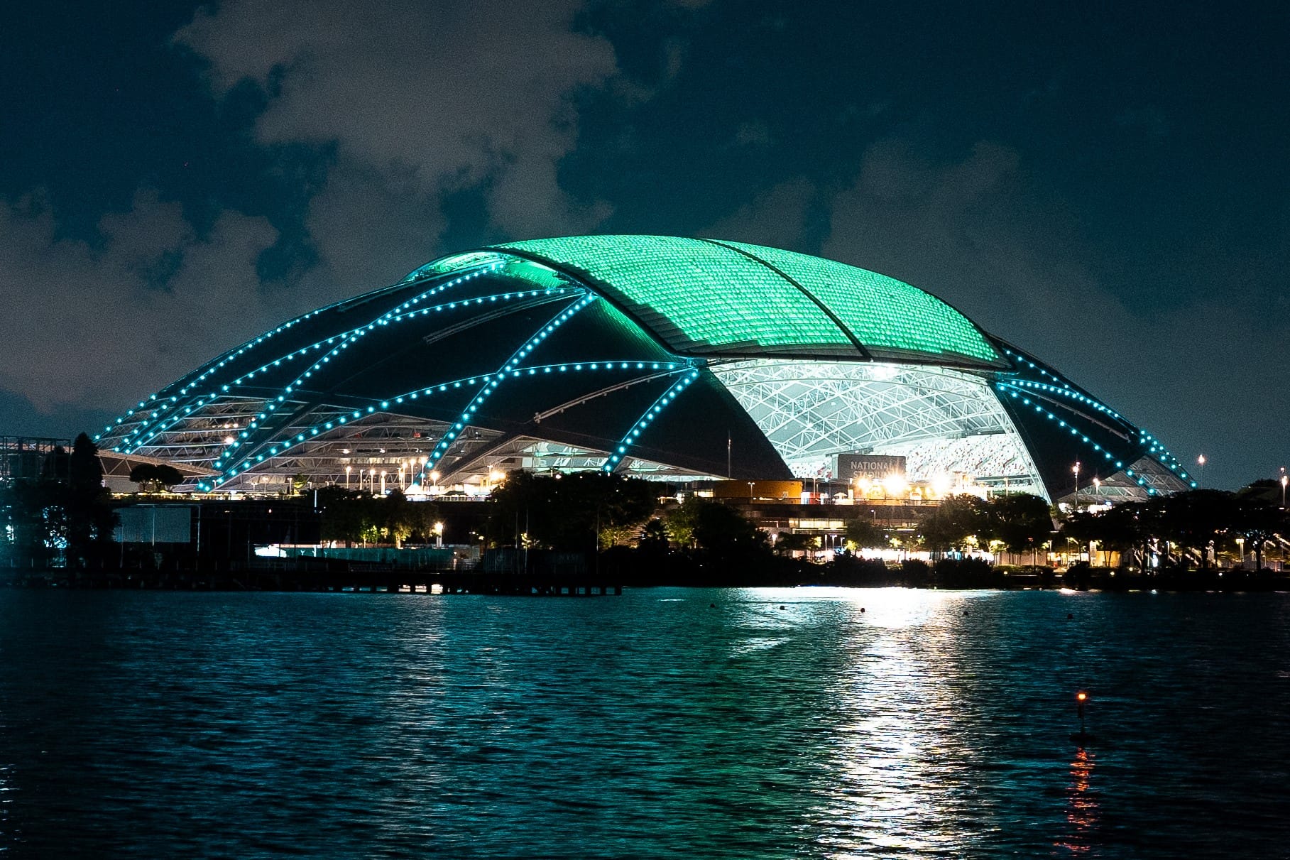 Lit-up National Stadium against a dark sky, reflected in water.