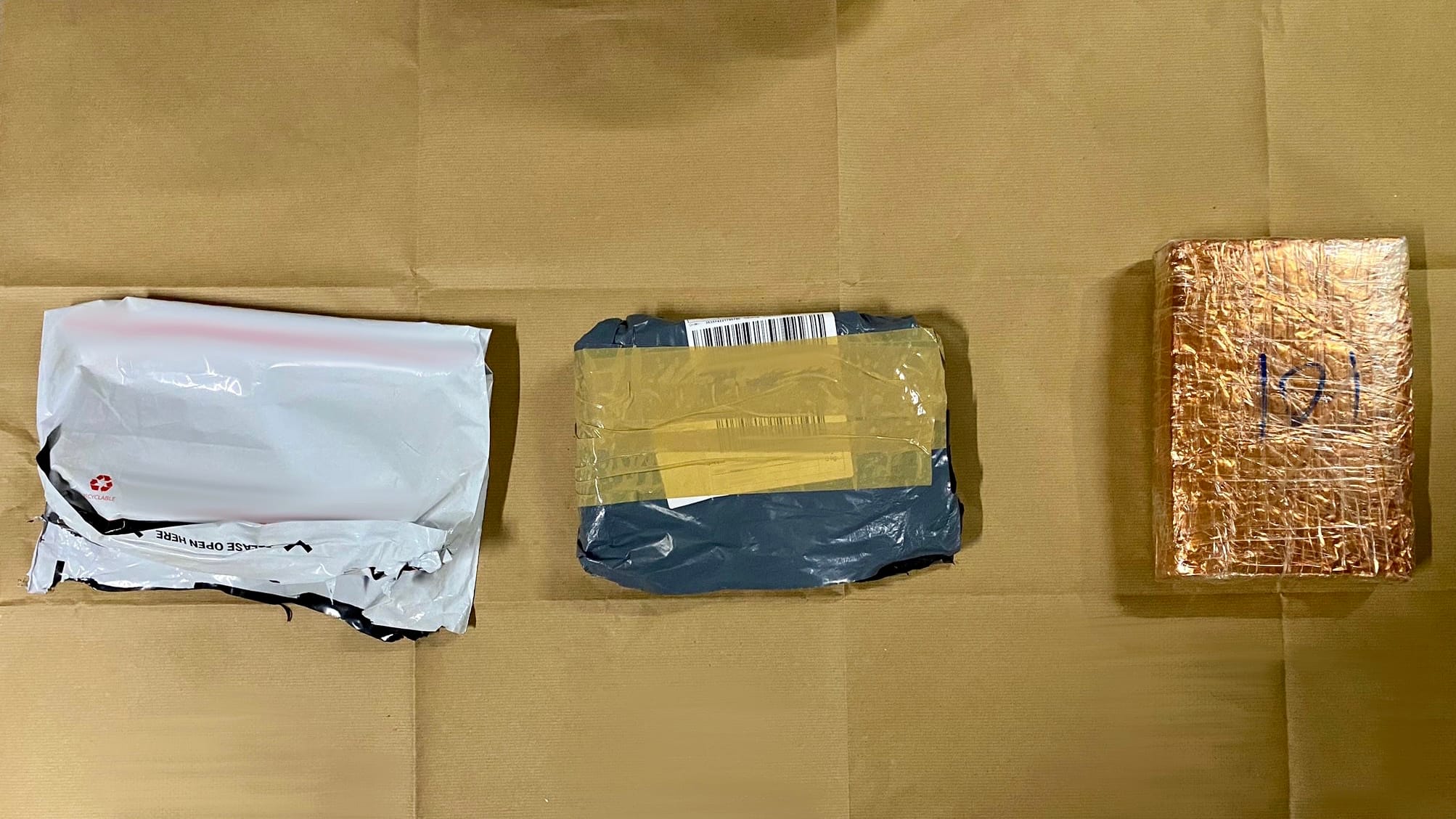 Three packages on brown paper: white envelope, blue taped box, foil-wrapped brick with writing.