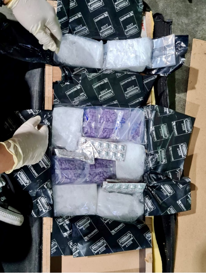 Gloved hands pack clear bags of white powder and purple pills inside a black Graphic 200T-lined container.