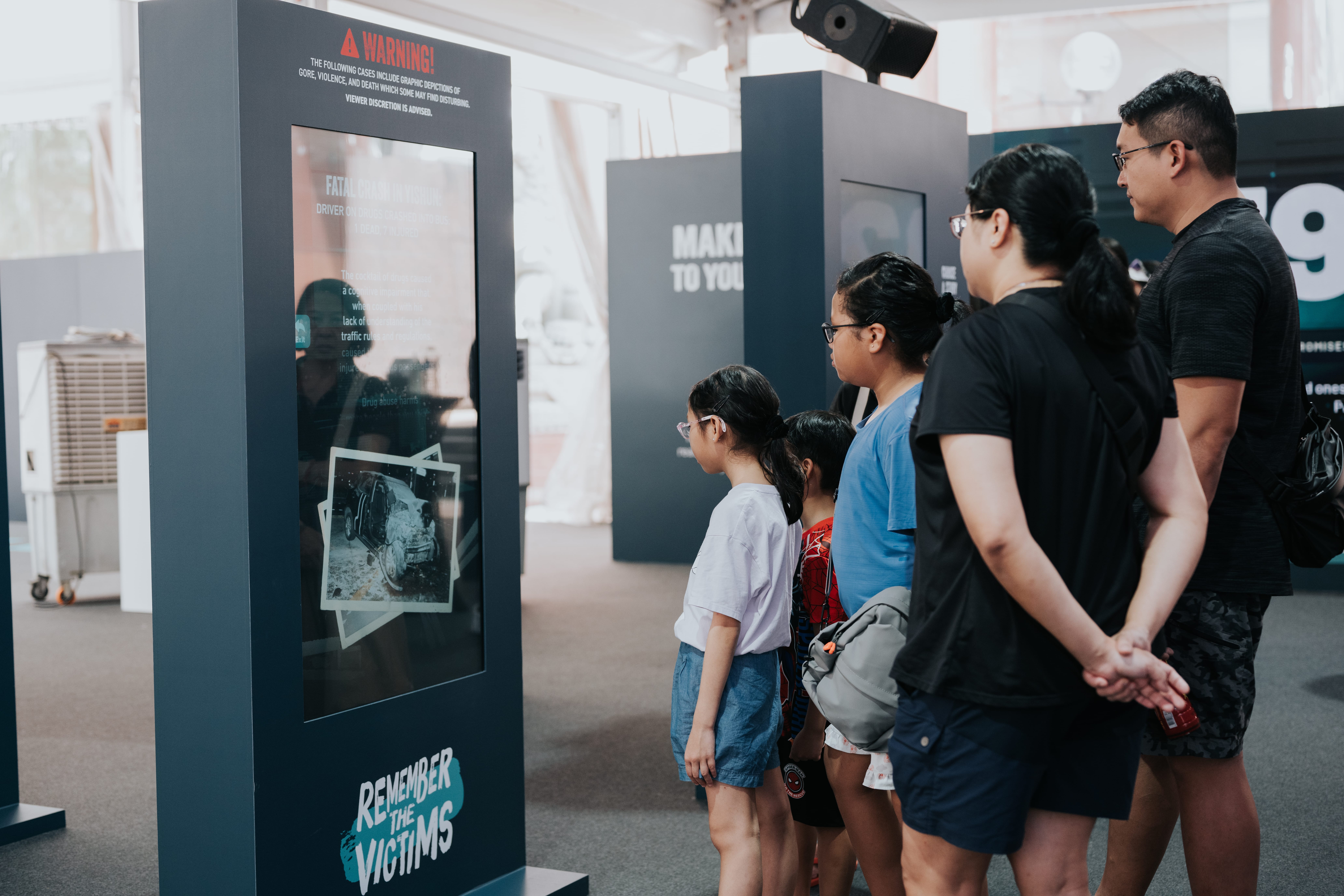 Family looking at 'Remember the Victims' exhibit with car crash photos and drug use warning.