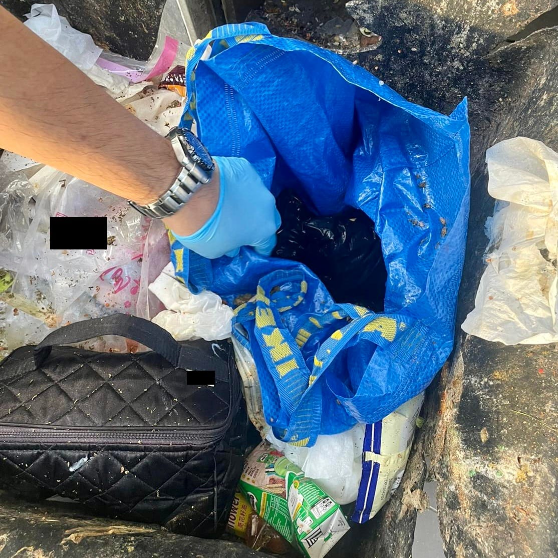 Gloved hand reaches into a blue IKEA bag inside a trash can full of refuse. A black bag sits within.
