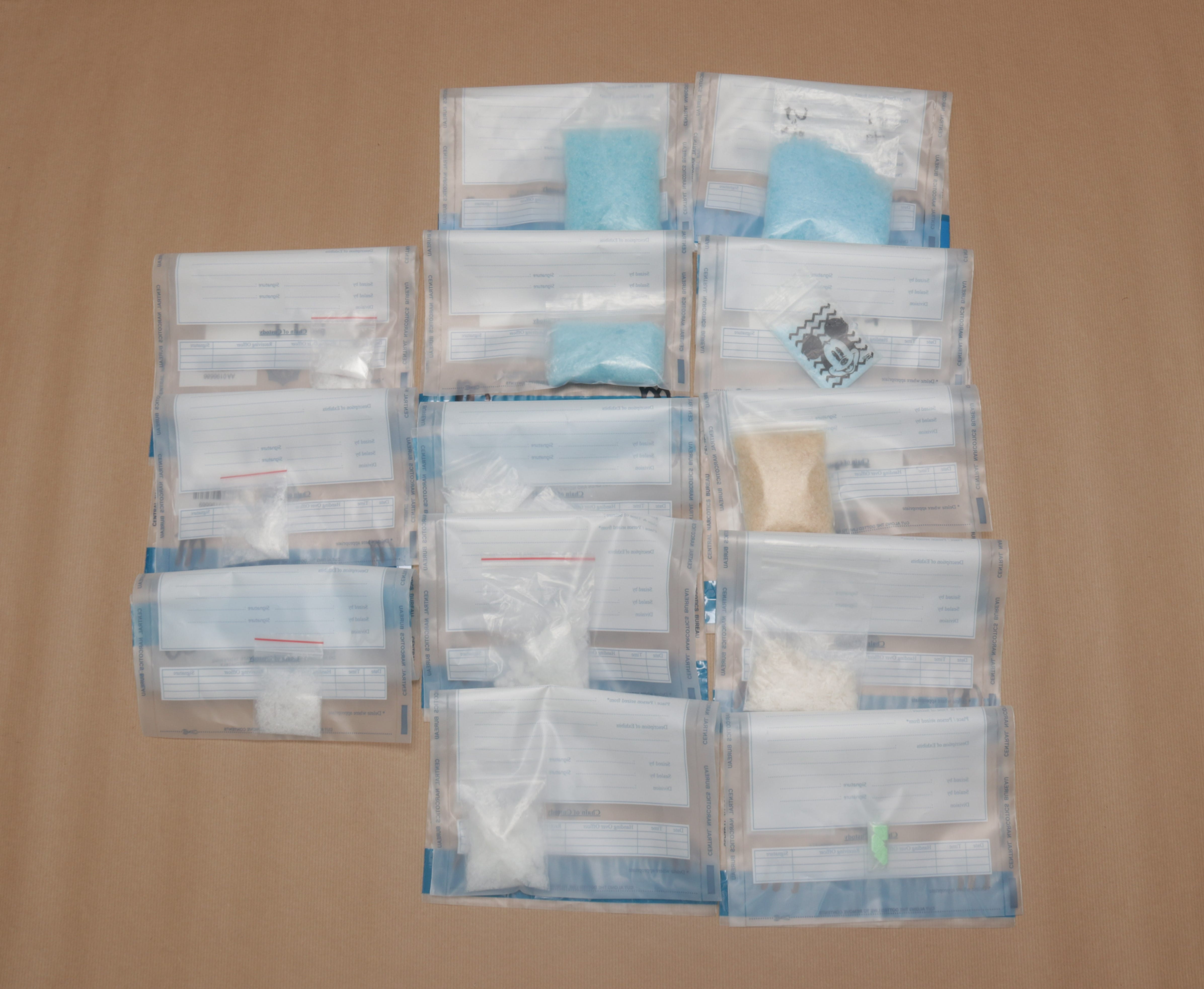 Clear evidence bags with blue, white, tan substances, and a Mickey Mouse pill, against a tan backdrop.