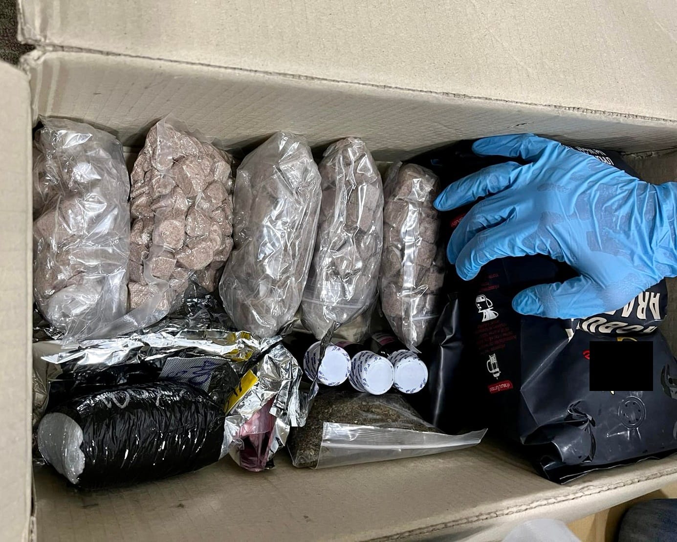 Box with plastic bags of brown disc-shaped items, loose substance, and black coffee bags. Blue-gloved hand reaches in.