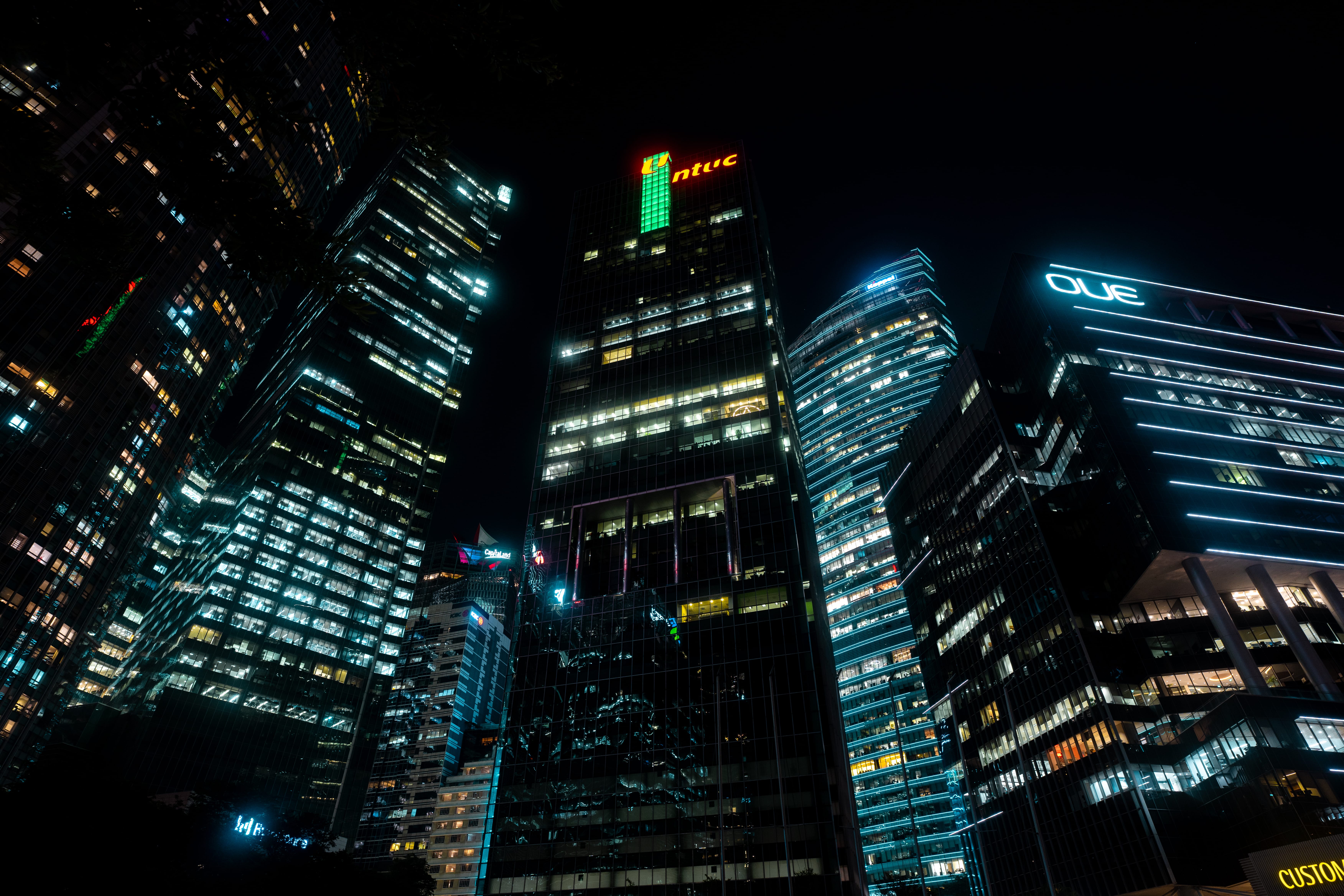 Skyscrapers at night with bright office windows, including buildings with NTUC and OUE signage.