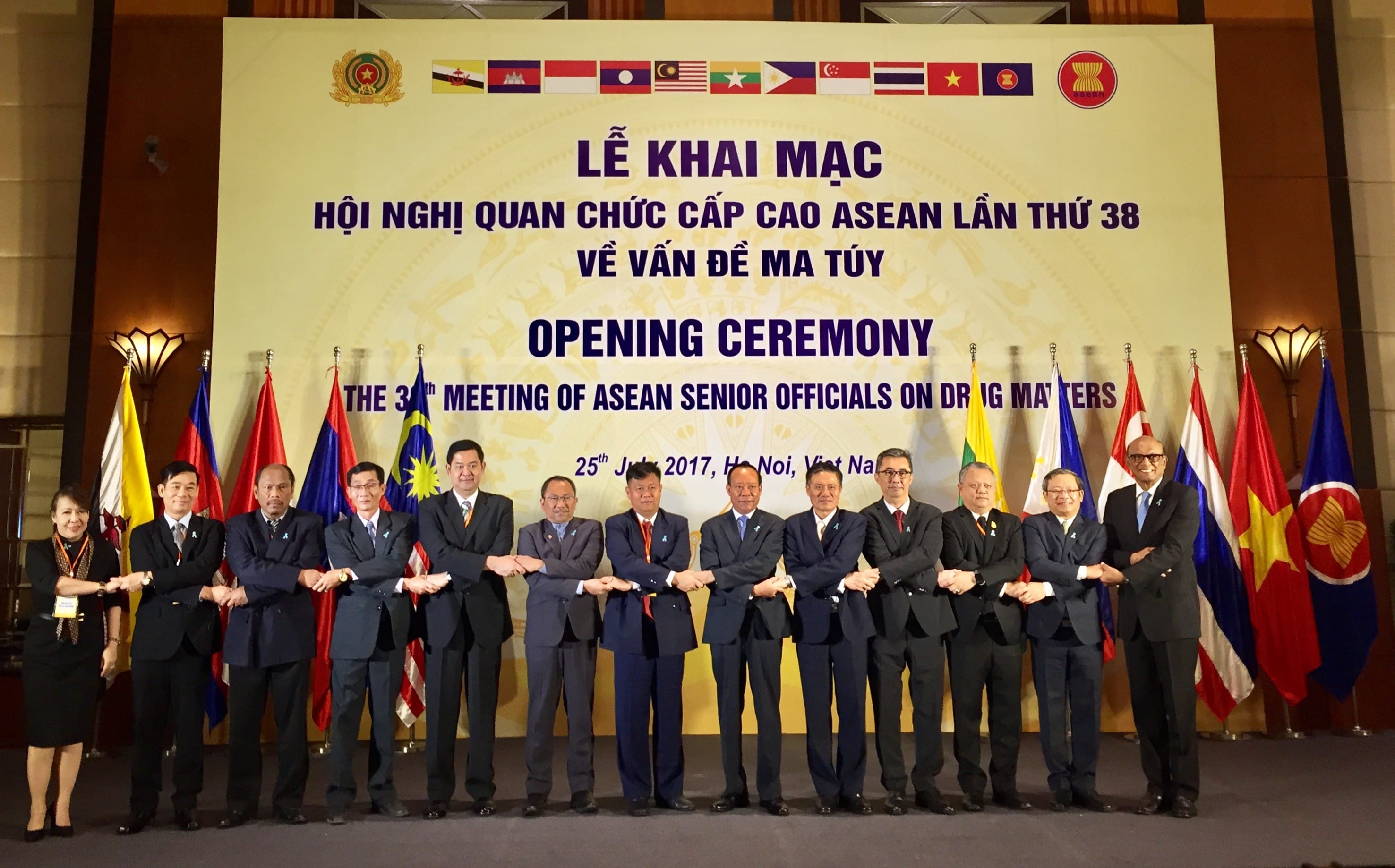 A group of dignitaries in suits link arms on stage before ASEAN flags and backdrop.