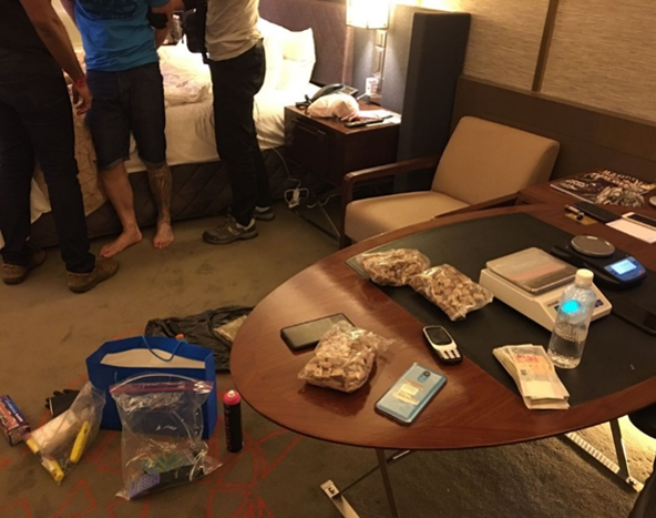 Hotel room with people, a desk laden with bagged material, scales, cash, and phones.