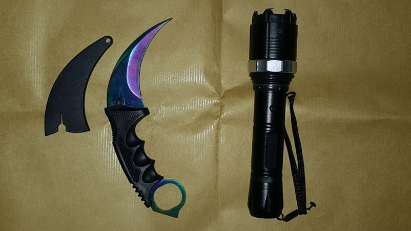 Karambit knife with sheath and black tactical flashlight on a brown surface.