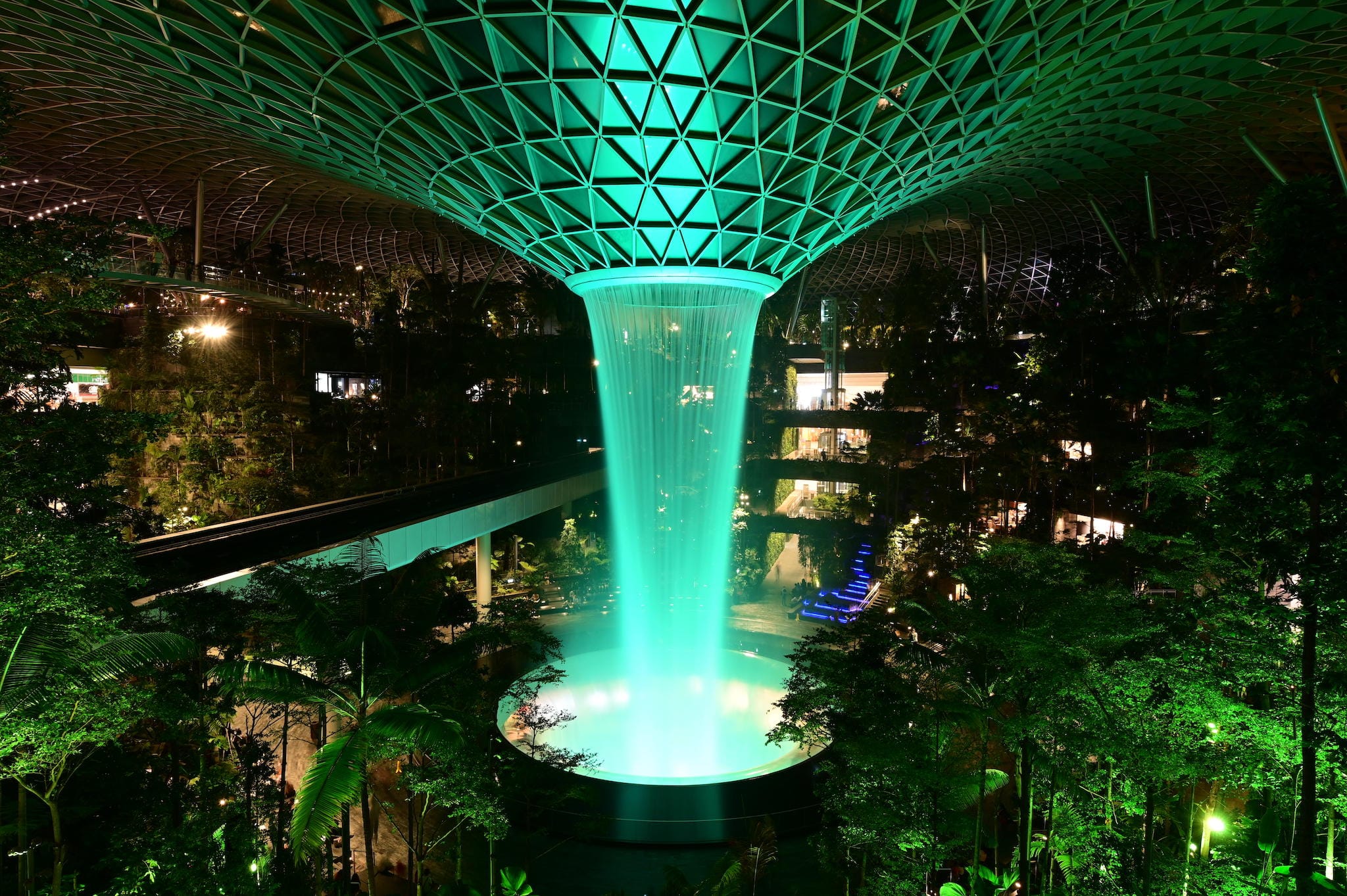 Indoor waterfall, glowing green, under geometric ceiling with plants around.