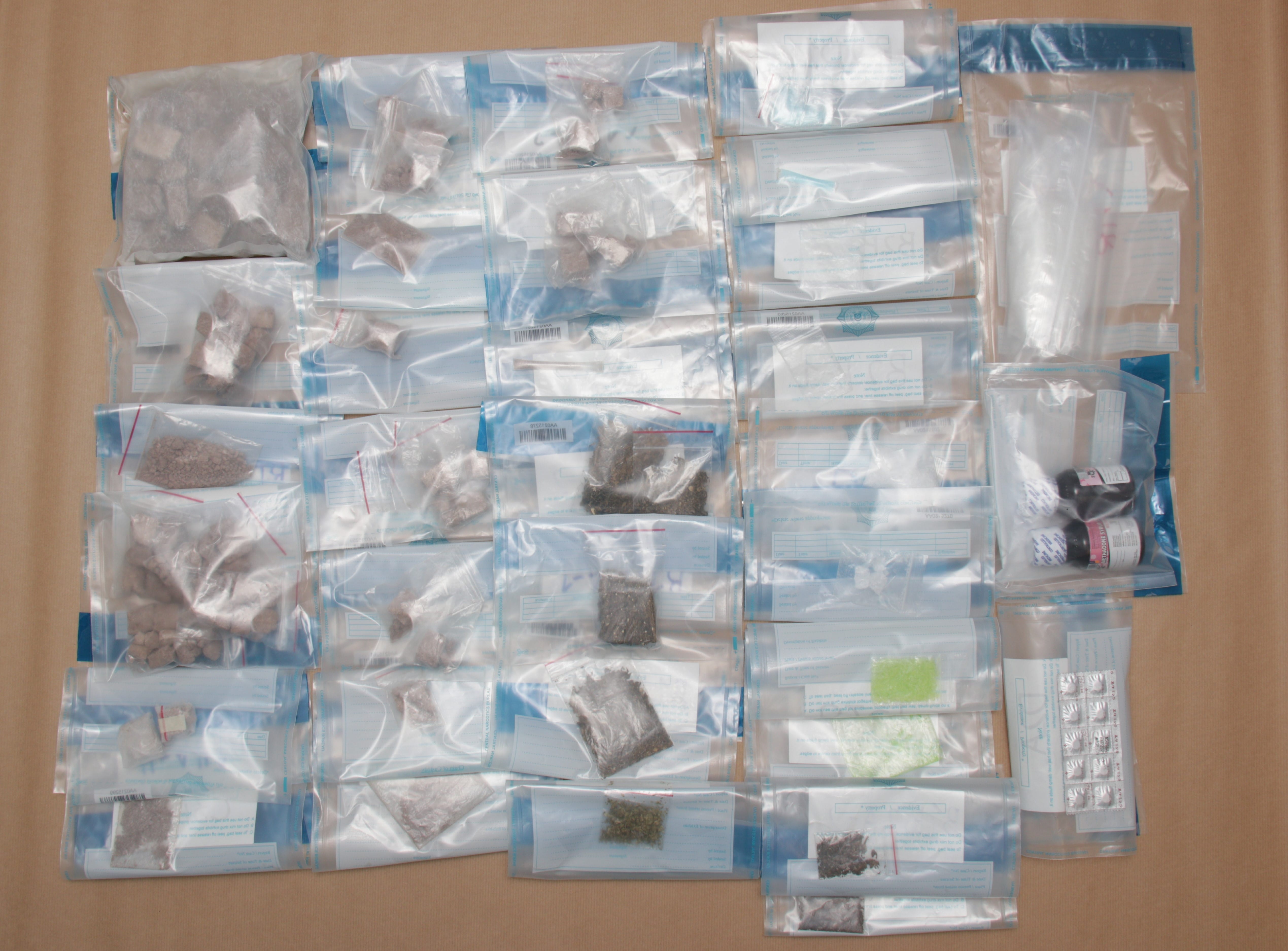Multiple clear plastic bags with contents (brown rocks, green leafy material, pills) on a brown surface.