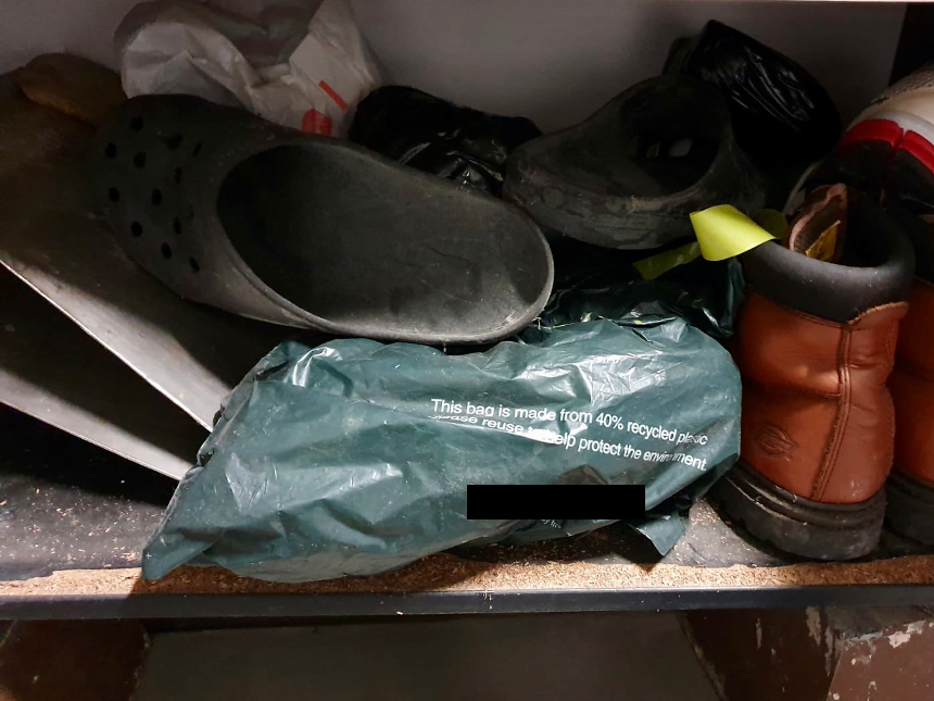 Shelf with assorted footwear, including black clogs, brown work boots, and a green recycled plastic bag.